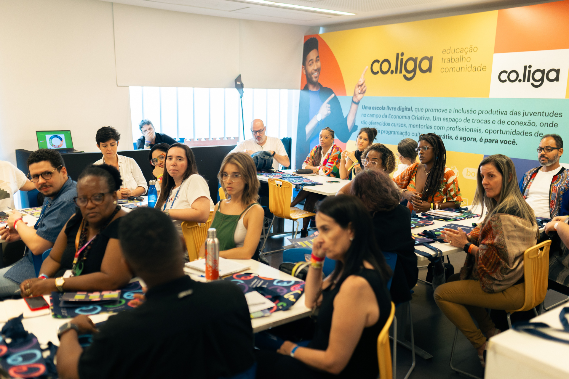 A foto mostra uma sala iluminada, com cerca de vinte pessoas sentadas em torno de mesas retangulares, participando de uma reunião ou oficina. Elas observam atentamente algo fora do quadro, algumas com cadernos e garrafas sobre as mesas. No fundo, há um grande painel colorido com a identidade visual da co.liga, que traz o texto: “educação, trabalho, comunidade” e uma breve descrição sobre o projeto. O ambiente é moderno e acolhedor, com cadeiras amarelas e clima de concentração e troca de ideias.