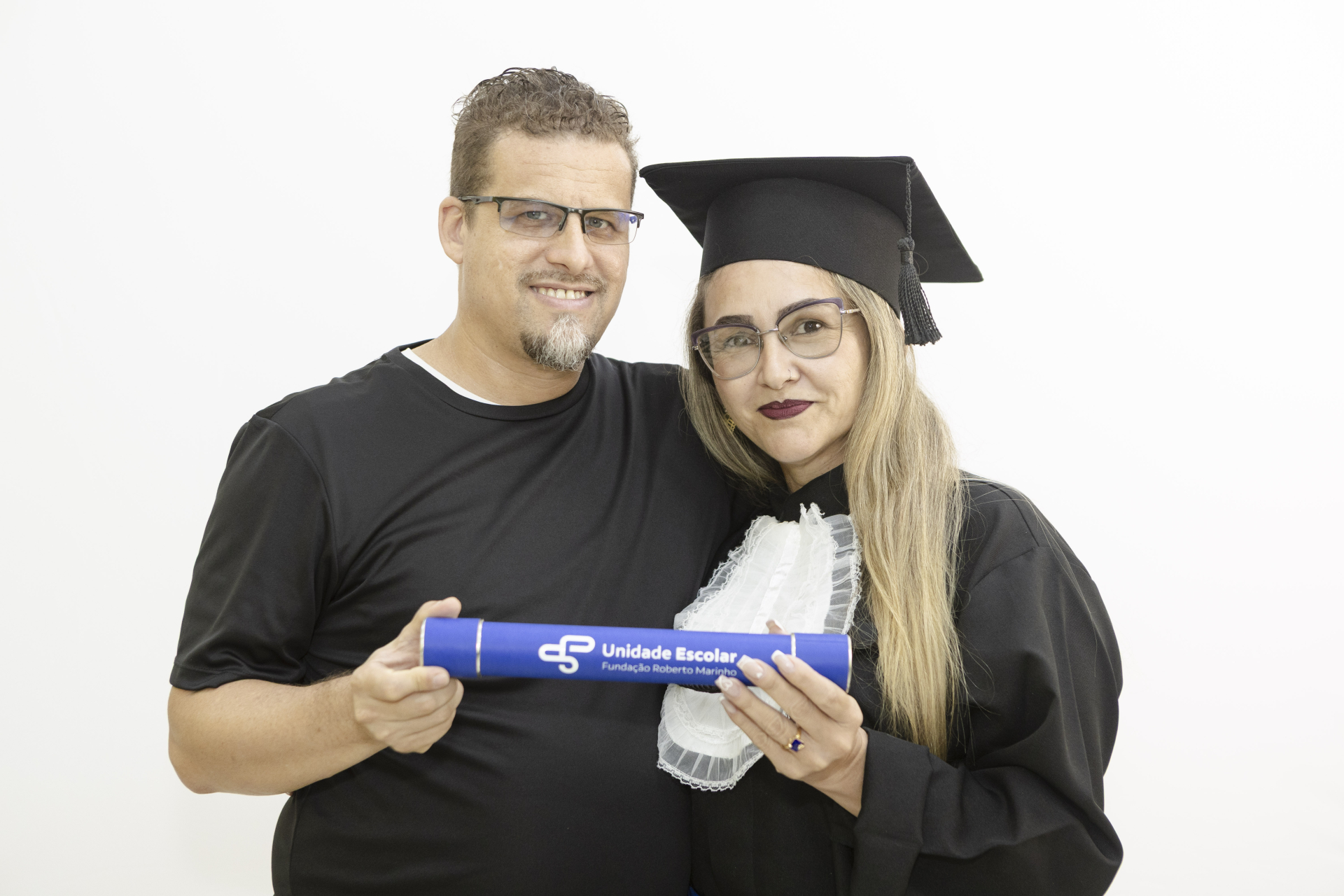 Um casal posando contra um fundo branco e liso. Uma mulher está à direita, vestindo uma beca de formatura preta com estola branca e um capelo. Ela tem cabelos longos e loiros, usa óculos e maquiagem escura nos lábios. Um homem está à esquerda, abraçando-a e sorrindo. Ele veste uma camiseta preta simples e usa óculos. Ambos seguram um diploma enrolado (canudo) de cor azul com a inscrição "Unidade Escolar Fundação Roberto Marinho" em destaque.