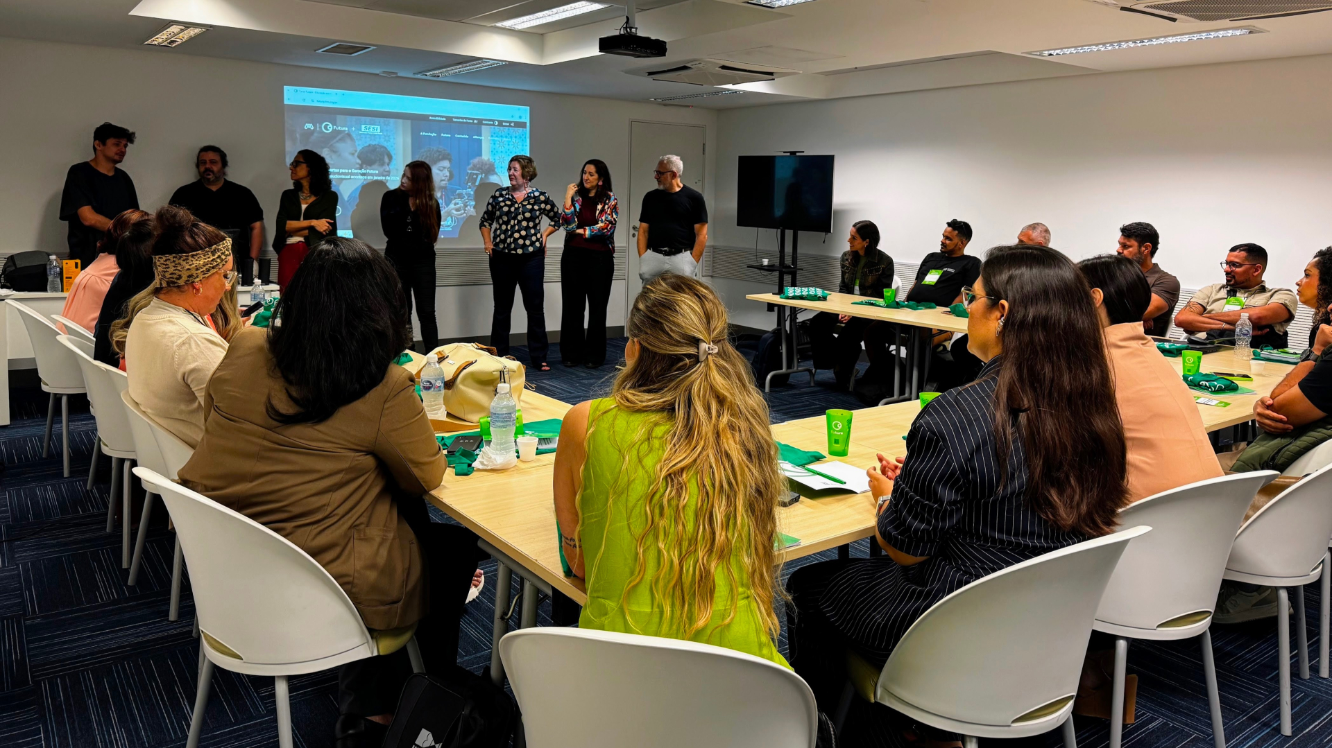Na imagem, um grupo de pessoas está reunido em uma sala de formação. Elas estão sentadas em mesas organizadas em formato de “U”, voltadas para a parte da frente da sala. No centro, de pé, há um pequeno grupo de facilitadores ou convidados apresentando algo ao público. Ao fundo, um projetor exibe uma imagem em uma tela na parede. Sobre as mesas, há materiais de apoio, copos verdes e garrafas de água. O clima é de atenção e troca, em um ambiente de encontro ou atividade colaborativa.