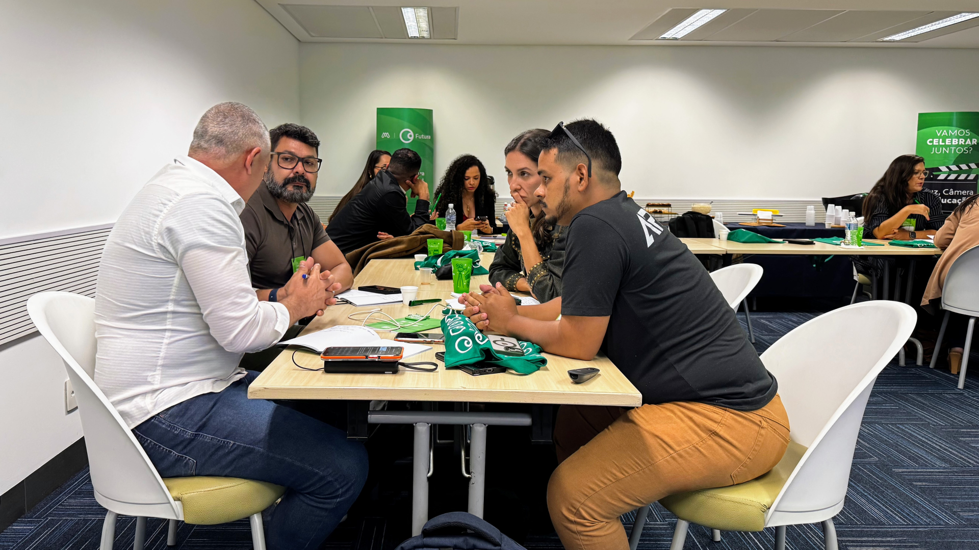 A imagem mostra um grupo de pessoas sentadas ao redor de uma mesa retangular, em um ambiente que parece ser uma sala de reunião ou formação. Quatro pessoas estão em destaque, conversando de forma concentrada. Elas estão inclinadas para a mesa, com expressão séria e atenta, sugerindo um diálogo ou trabalho colaborativo. Sobre a mesa há garrafas de água, copos verdes, cadernos, canetas, celulares e bolsas de tecido verdes com uma marca ou logotipo. Ao fundo, outras pessoas também estão reunidas em mesas, em c