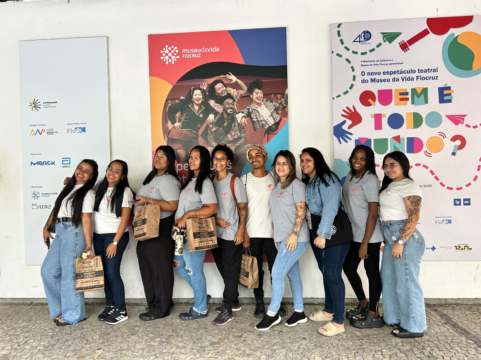 Um grupo de dez jovens, nove mulheres e um homem, todos vestindo camisetas cinzas uniformizadas com um pequeno emblema no peito, posam lado a lado em frente a uma parede com cartazes coloridos sobre eventos culturais e o Museu da Vida Fiocruz. O grupo está sorrindo. Muitos estão segurando sacolas de papel marrons nas mãos, provavelmente de brindes ou compras. O jovem no centro se destaca com um gorro e é o único homem do grupo. O cenário sugere um passeio ou visita a um local cultural.
