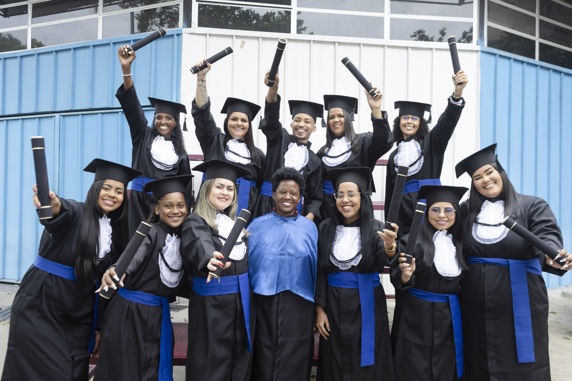 Um grupo de dez mulheres sorridentes, todas vestindo becas de formatura pretas com estolas brancas e faixas azuis na cintura. Elas também usam o capelo preto de formatura. A maioria segura o diploma enrolado (canudo) para o alto em um gesto de celebração. No centro do grupo, uma mulher, que parece ser a professora ou coordenadora, está vestida com uma camisa azul-claro vibrante e calças pretas, em contraste com as formandas. O fundo é uma parede externa de metal em tons de branco e azul. A foto transmite al