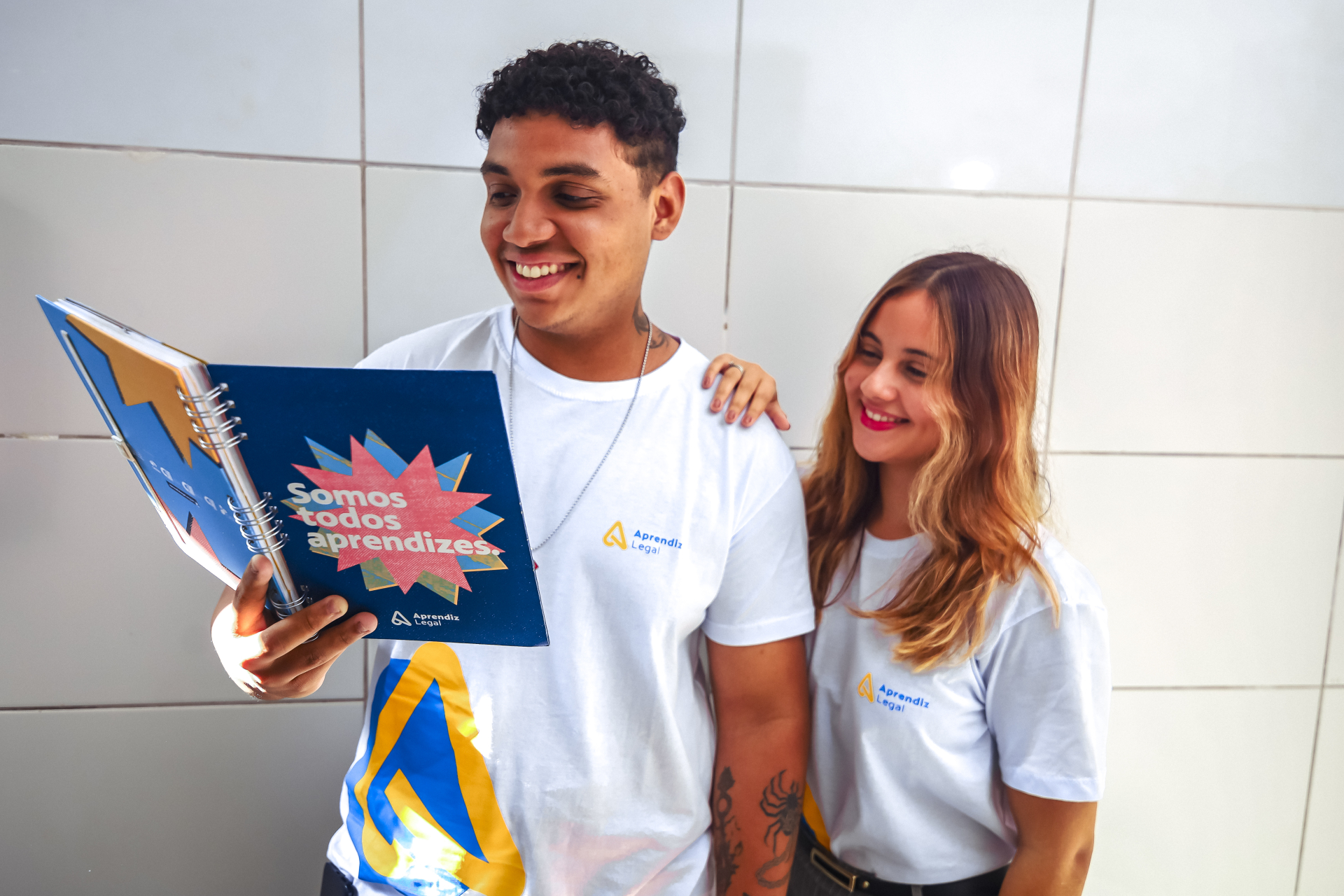 Duas pessoas jovens estão de pé, lado a lado, em frente a uma parede clara revestida de azulejos. Ambas vestem camisetas brancas com o logotipo “Aprendiz Legal” estampado no peito e em tamanho maior na parte inferior da camisa. A pessoa à esquerda segura um caderno espiral azul com a frase em destaque: “Somos todos aprendizes”, escrita dentro de uma forma colorida semelhante a uma explosão gráfica. Ela sorri enquanto olha para o caderno. Tem cabelo curto e escuro. A pessoa à direita também sorri e observa