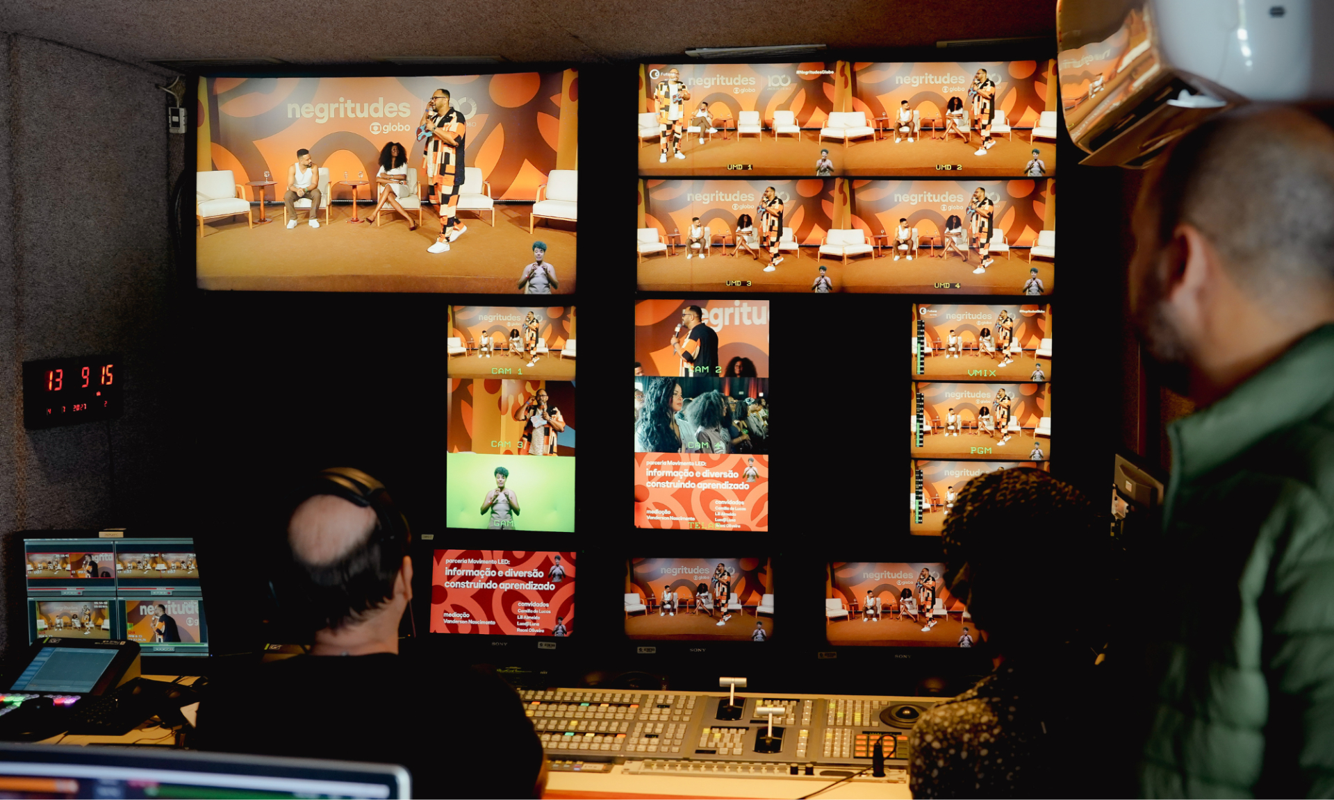 Sala de controle de TV ou estúdio de transmissão. Várias telas mostram diferentes enquadramentos de um evento chamado “negritudes”, com pessoas no palco falando. À frente das telas, técnicos observam e operam equipamentos. O ambiente é escuro, iluminado principalmente pelos monitores.