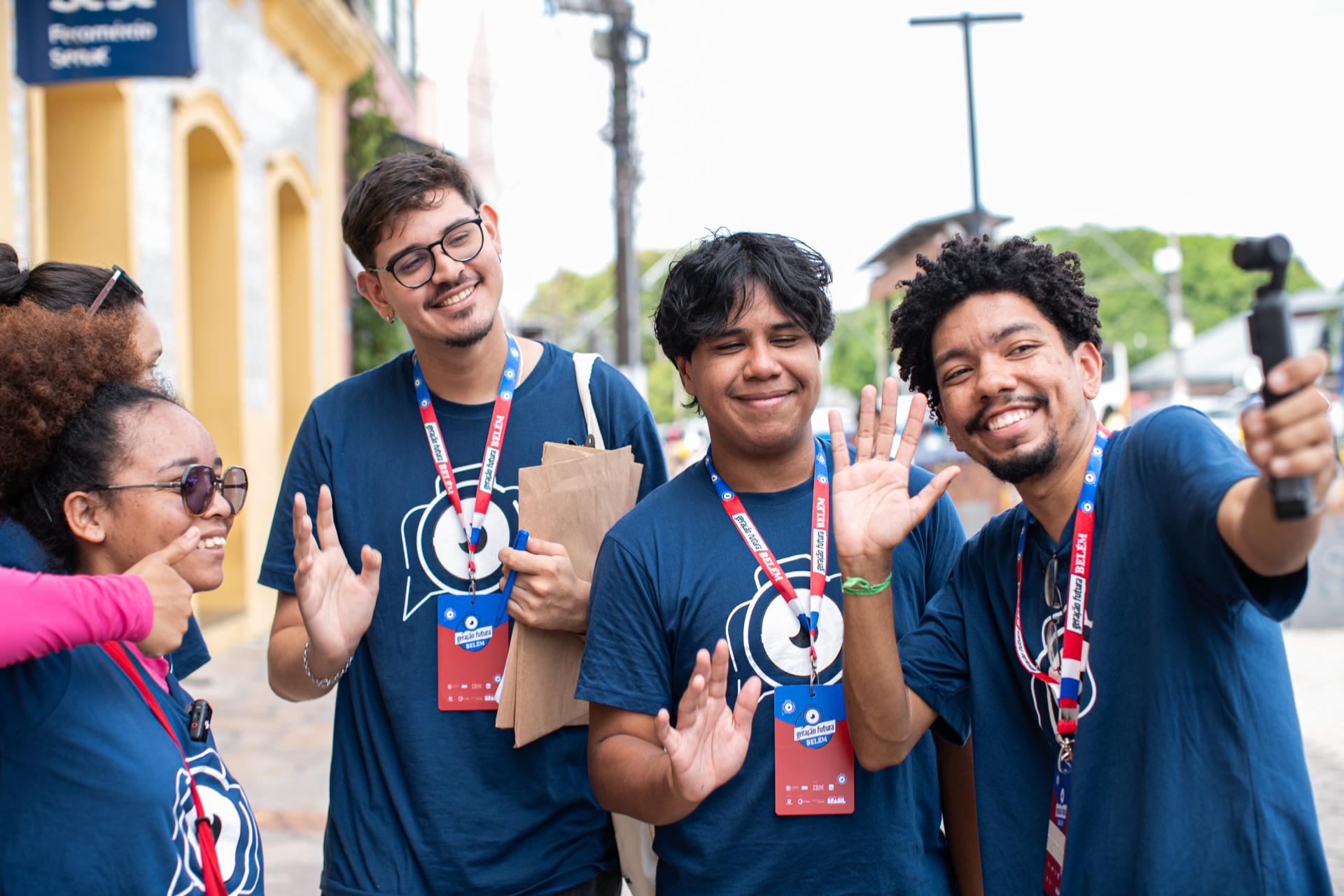 Um grupo de jovens usa camisetas azuis e crachás de identificação enquanto interage de forma descontraída em um espaço urbano. Eles sorriem, acenam para a câmera e um dos integrantes registra o momento com um celular acoplado a um estabilizador, sugerindo a produção de conteúdo ou a cobertura de um evento.