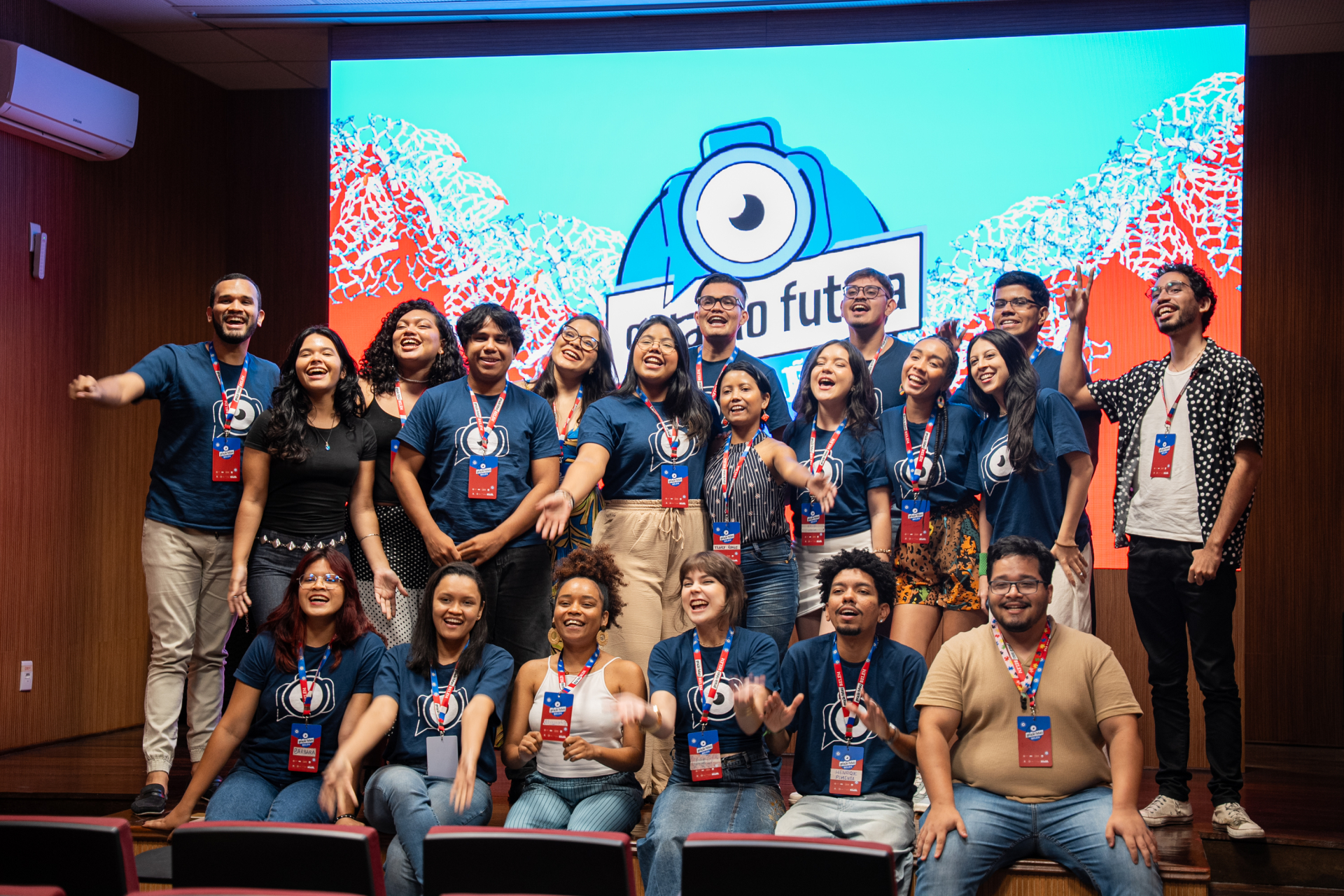 Grupo de jovens animados, com os braços esticados e sorrindo. Eles vestem camisetas azul escuro, com o logo do Geração Futura. O grupo, com cerca de 20 pessoas, está a frente de um telão iluminado.