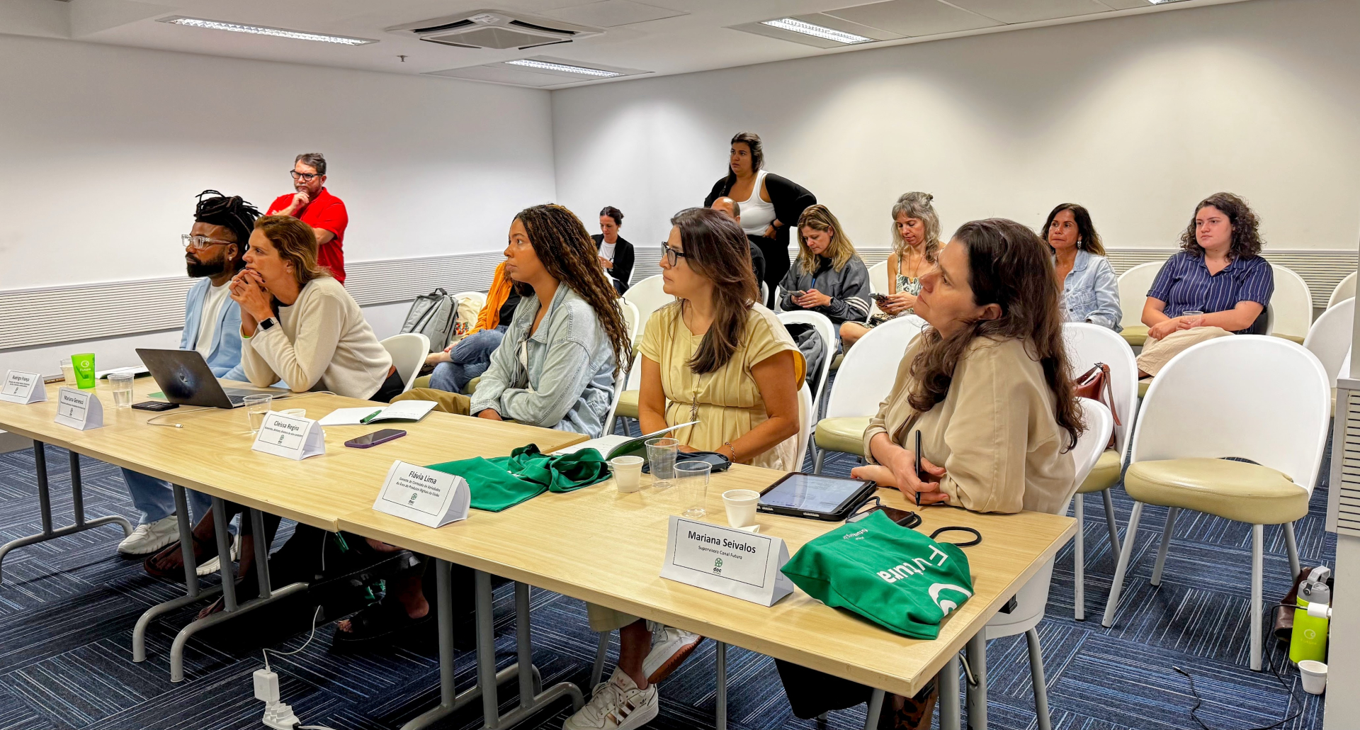 A foto mostra uma sala de reuniões com várias pessoas sentadas, prestando atenção a algo fora do enquadramento, à esquerda. Na parte da frente, há uma mesa comprida com cinco pessoas sentadas lado a lado. Atrás desse grupo, outras pessoas estão sentadas em cadeiras brancas, formando uma segunda fileira. Elas também observam a atividade com atenção; algumas usam celulares ou seguram objetos no colo. 