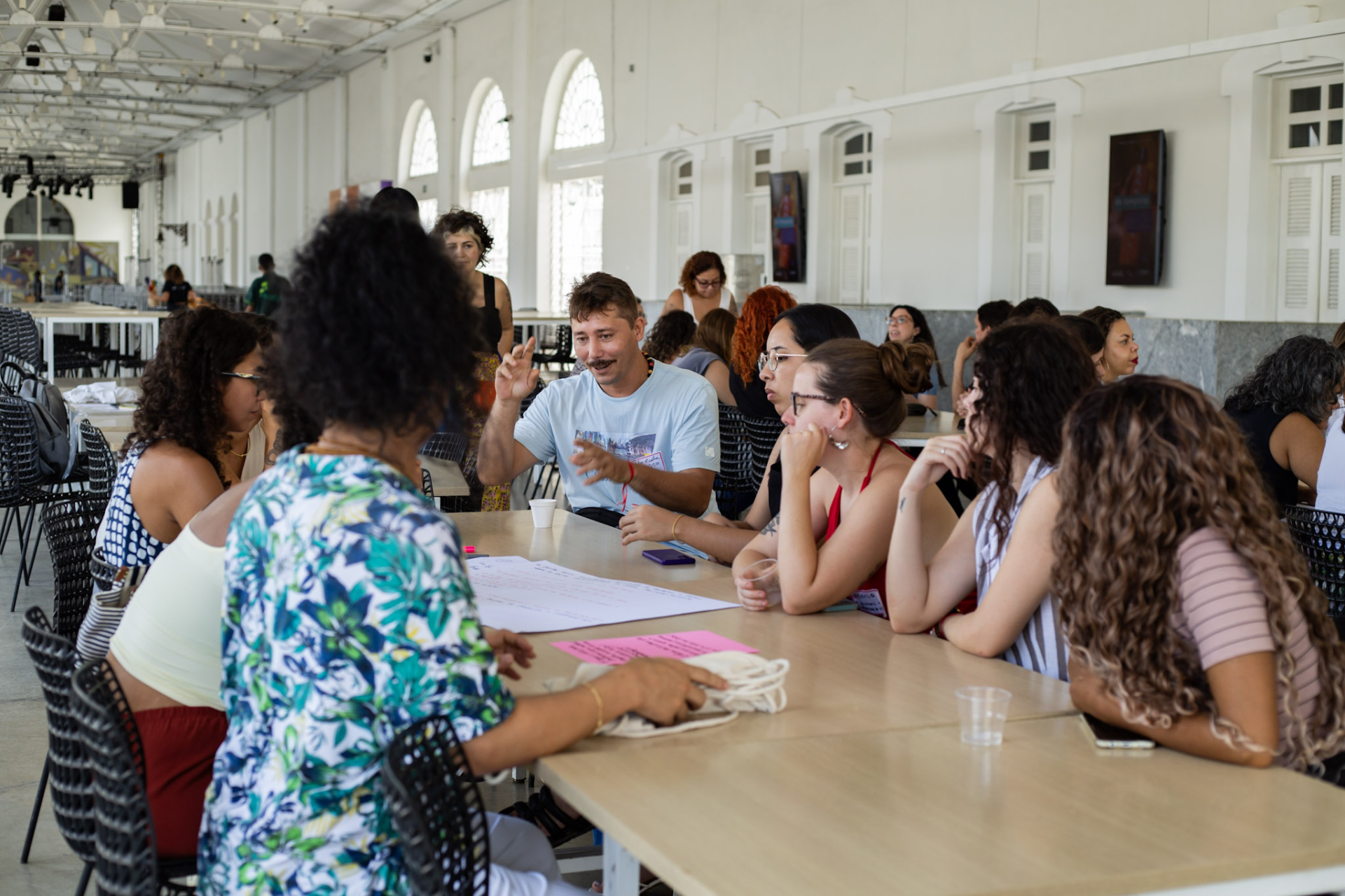 A imagem mostra um grupo de cerca de dez pessoas sentadas ao redor de uma mesa retangular de madeira clara, em um grande salão amplo e iluminado. O espaço tem teto alto, estruturas metálicas aparentes e várias janelas grandes em arco ao longo da parede, por onde entra bastante luz natural. Ao fundo, há outros grupos de pessoas sentados em mesas semelhantes, participando de atividades ao mesmo tempo.