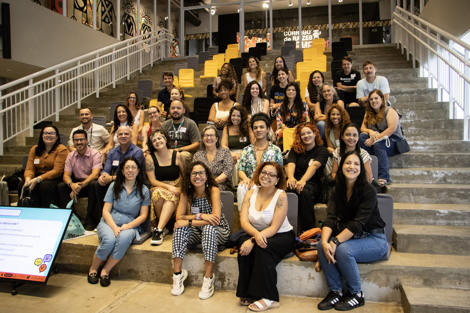 Fotografia em ambiente interno de um auditório com arquibancadas de concreto e assentos em amarelo e cinza. Aproximadamente 40 pessoas estão sentadas em fileiras, todas voltadas para a câmera e sorrindo. O grupo é diverso em gênero, idade e características físicas. As pessoas usam roupas casuais e profissionais, como vestidos, blusas, camisetas, calças jeans e tênis. Algumas usam crachás. Na parte inferior esquerda da imagem, aparece parcialmente um monitor com uma apresentação exibida. Ao fundo, há paredes