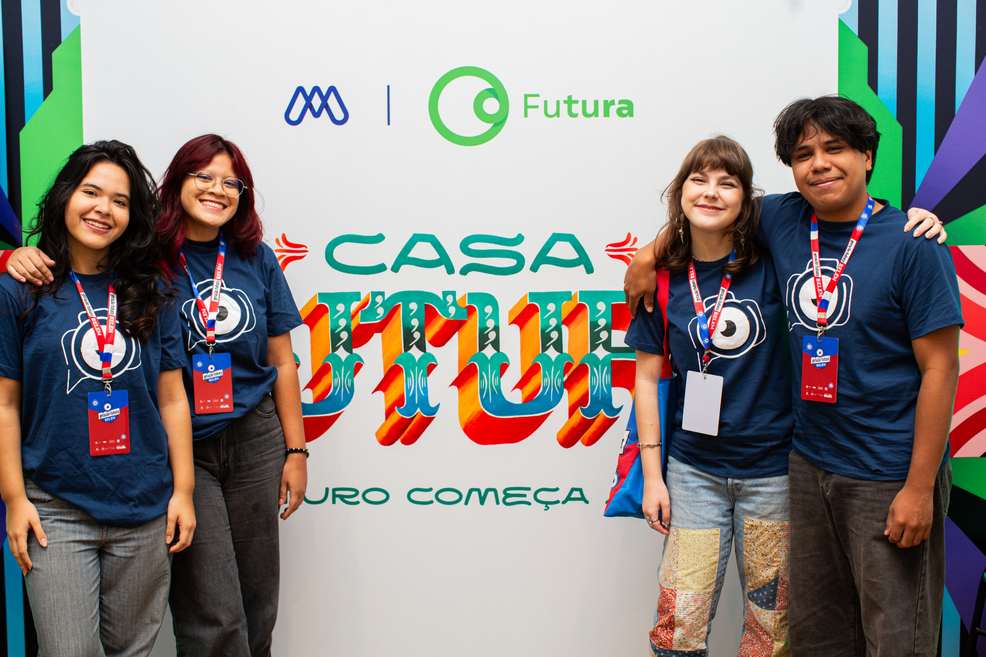 Quatro jovens posam sorridentes diante de um painel colorido com a identidade visual da Casa Futura. Eles vestem camisetas azuis com o mesmo símbolo gráfico, usam crachás de identificação e estão lado a lado, com braços apoiados nos ombros uns dos outros, demonstrando proximidade e espírito de equipe. Ao fundo, aparecem os logotipos institucionais e a frase “Casa Futura”, em letras estilizadas e multicoloridas.