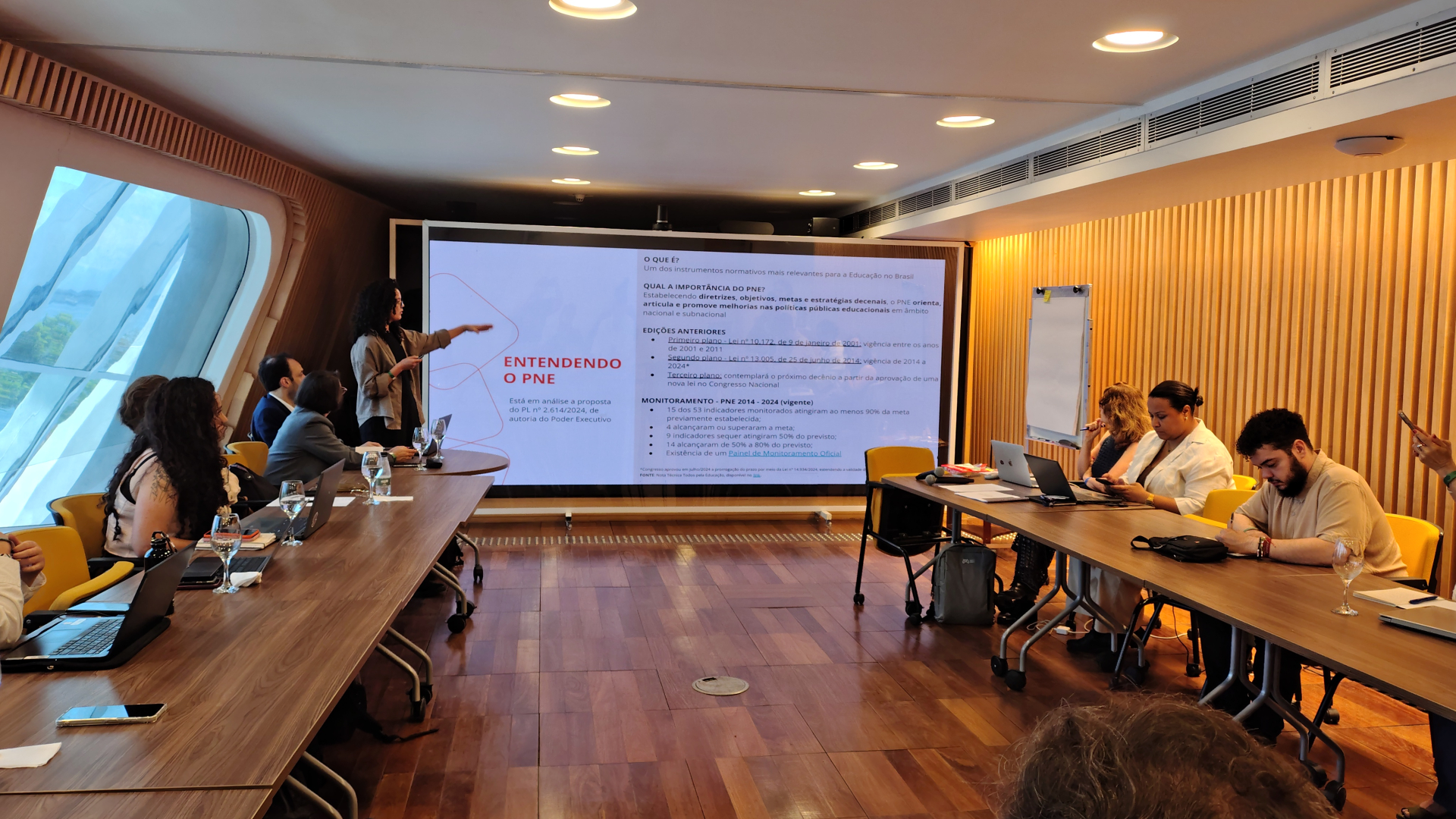 Sala de reunião comprida, com piso de madeira e paredes claras. À esquerda, uma mulher de cabelos cacheados, em pé ao lado de um telão grande, aponta para o slide projetado com o título em vermelho “ENTENDENDO O PNE” e texto em parágrafos. Em torno de uma mesa em U há várias pessoas sentadas, olhando para a apresentação, usando notebooks ou fazendo anotações; algumas taças de água estão sobre a mesa. À direita, a parede é revestida por ripas de madeira verticais e há um quadro branco com cavalete encostado.