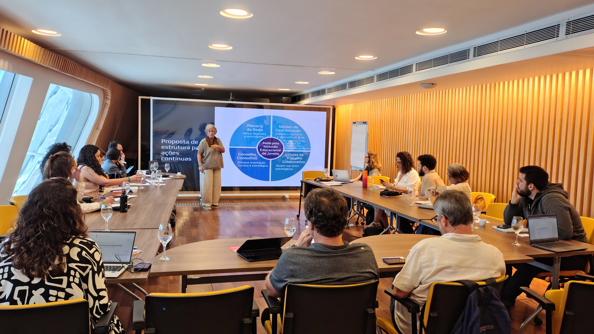 Reunião de trabalho com especialistas reunidos em uma sala iluminada, acompanhando a apresentação de uma palestrante diante de uma tela com a proposta de estrutura para ações contínuas. Participantes analisam materiais, fazem anotações e interagem, em um ambiente colaborativo e focado em planejamento estratégico.