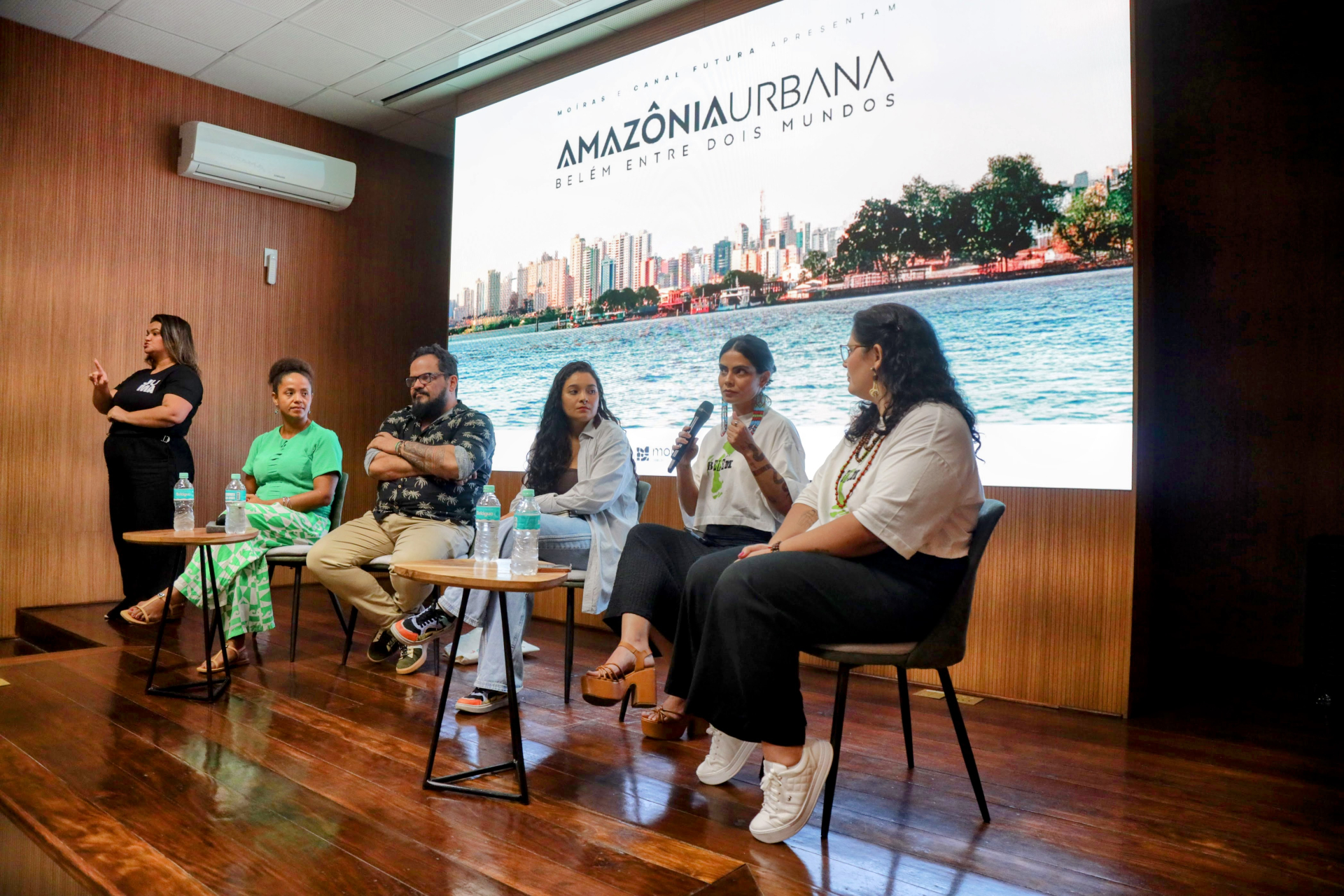Cinco pessoas sentadas em frente a um telão iluminado. Na TV, a uma imagem de paisagem com o título Amazônia Urbana. As pessoas sentadas parecem estar falando e algumas seguram o microfone.