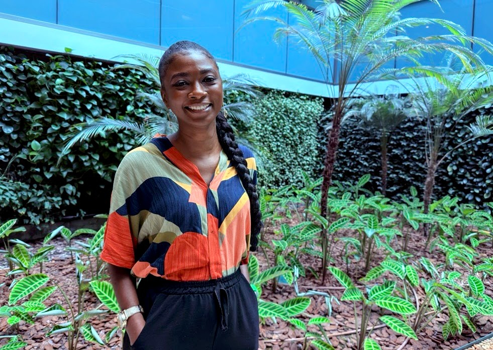 Uma mulher jovem está em pé em um jardim com muitas plantas verdes e palmeiras baixas. Ela sorri para a câmera e usa uma camisa colorida em tons de laranja, azul, verde e preto, combinada com calça preta. Seu cabelo está preso em uma longa trança lateral. Atrás dela, há paredes cobertas por plantas e um prédio com fachada azul. A imagem passa uma sensação de tranquilidade e acolhimento.