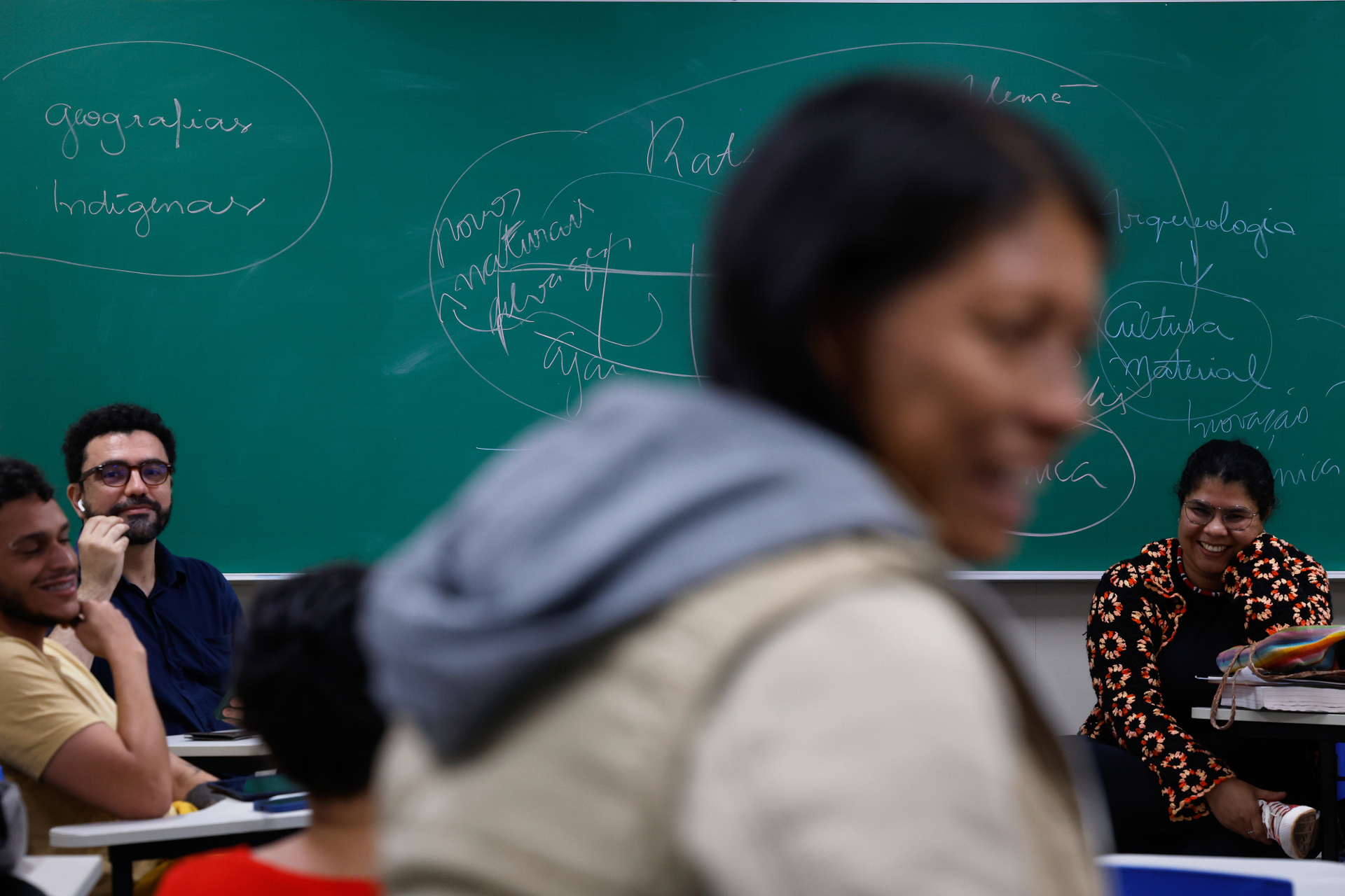 A imagem mostra uma sala de aula com pessoas sentadas, conversando e sorrindo. Ao fundo, há um quadro verde cheio de anotações e círculos feitos com giz. Em primeiro plano, uma pessoa aparece desfocada passando pela frente. A atmosfera é descontraída e parece haver uma aula ou discussão em andamento.