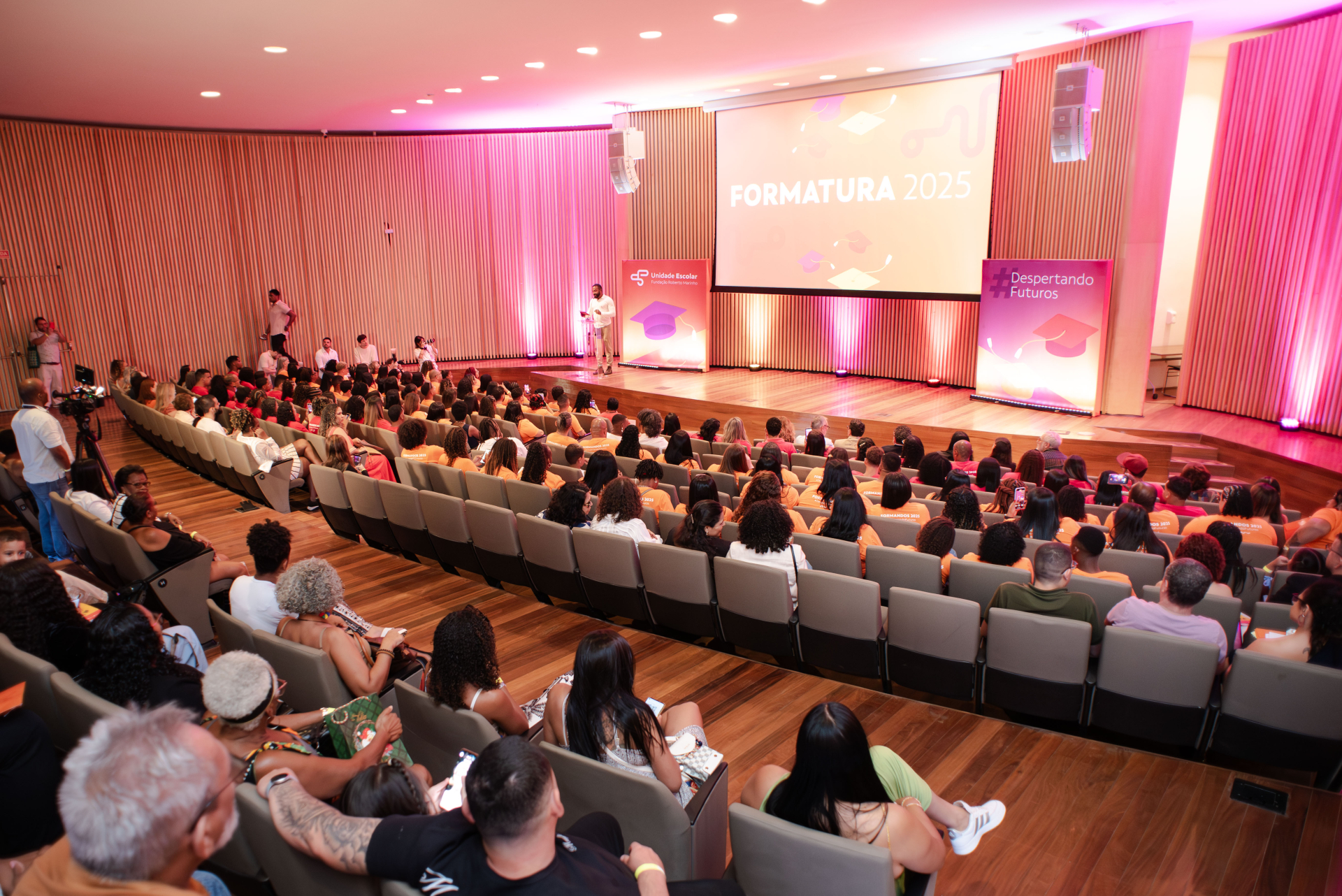 Auditório lotado durante a cerimônia de formatura de 2025, com o público atento ao palco enquanto um apresentador conduz o evento. A iluminação em tons de rosa e roxo e os painéis temáticos reforçam o clima de celebração e conquista