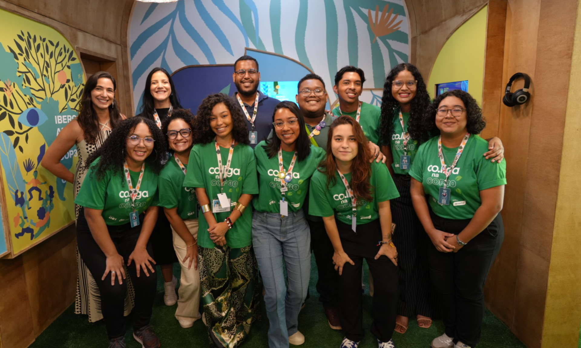 Foto de grupo com cerca de 13 pessoas, em sua maioria jovens adultos, posando lado a lado e sorrindo para a câmera. A maioria veste camisetas verdes com a inscrição “co.liga na COP30” e usa crachás pendurados no pescoço. O grupo está em um espaço interno decorado, com paredes em tons de bege e painéis coloridos ao fundo, com ilustrações de folhas, plantas e figuras humanas. O ambiente remete a um estande ou área institucional de evento.