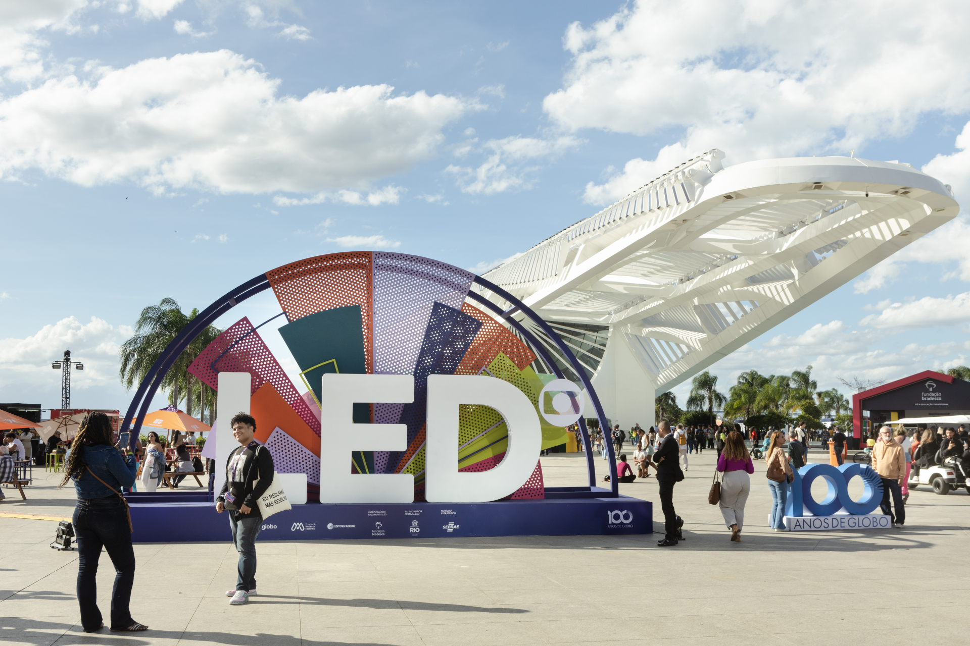 Em uma grande praça ao ar livre, durante o dia, há uma instalação colorida com as letras grandes “LED” em branco, apoiadas sobre uma base azul. Atrás das letras, um arco semicircular é formado por painéis coloridos e perfurados, em tons de laranja, roxo, verde e azul. Ao redor da instalação, várias pessoas caminham, conversam ou tiram fotos; algumas estão paradas observando o espaço.