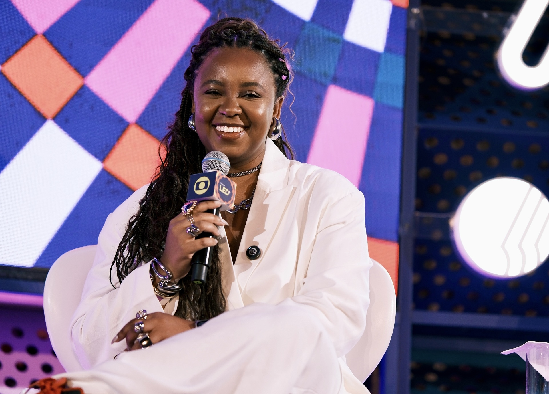 A imagem mostra uma mulher sentada em um palco, sorrindo enquanto fala ao microfone em um evento público. Ela veste um traje claro, elegante, e usa acessórios como anéis, pulseiras e brincos, que reforçam um visual sofisticado e confiante. O microfone tem um identificador de mídia, sugerindo que se trata de uma conversa, painel ou entrevista mediada.