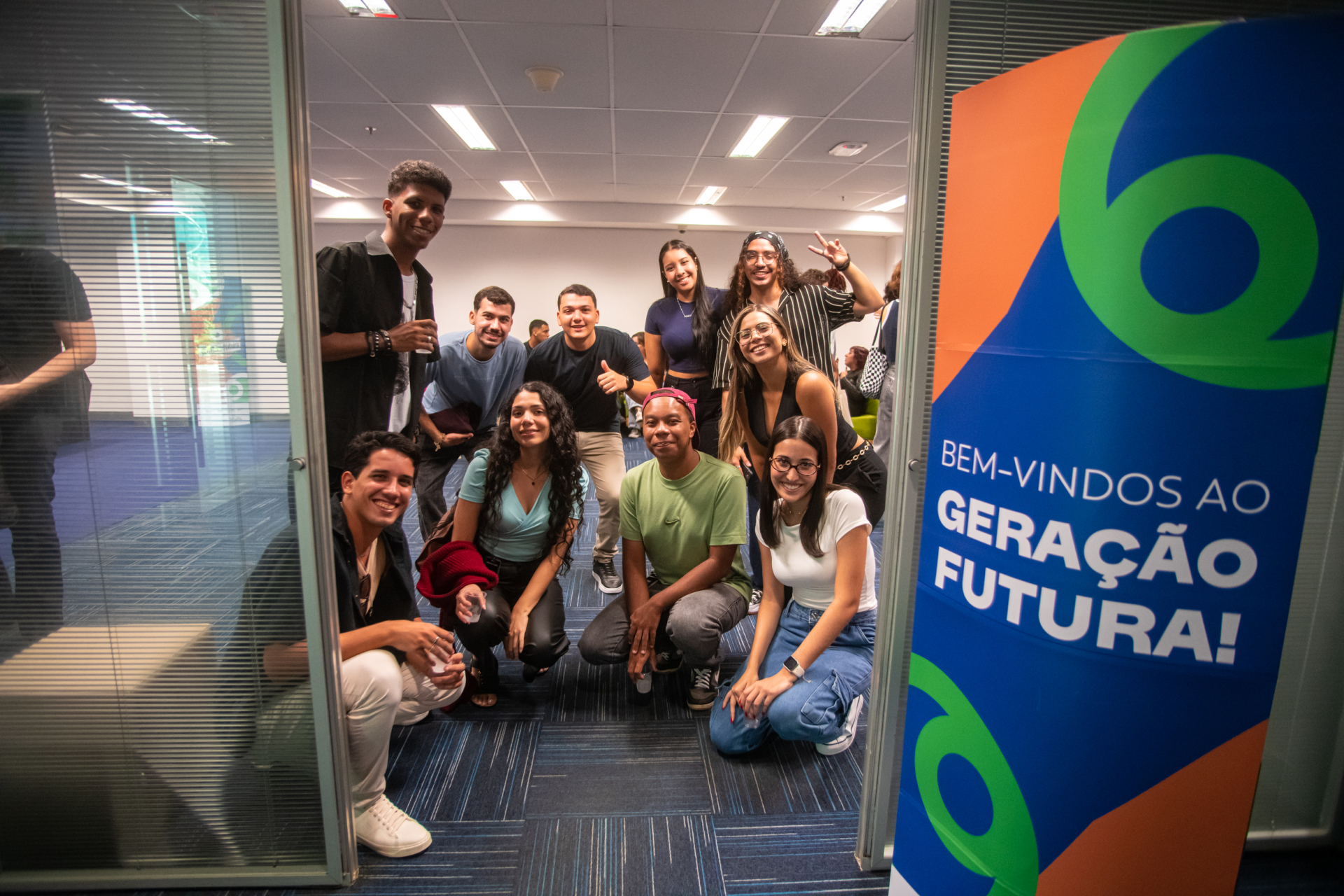 Um grupo de jovens posa sorridente dentro de uma sala de eventos, reunido em frente à porta de entrada, em um ambiente corporativo iluminado. Alguns estão agachados e outros em pé, demonstrando descontração e espírito de grupo. À direita da imagem, um painel colorido traz a mensagem “Bem-vindos ao Geração Futura!”, reforçando o caráter de acolhimento e formação do encontro. O cenário e as expressões dos participantes sugerem um momento de integração, celebração e troca entre jovens.