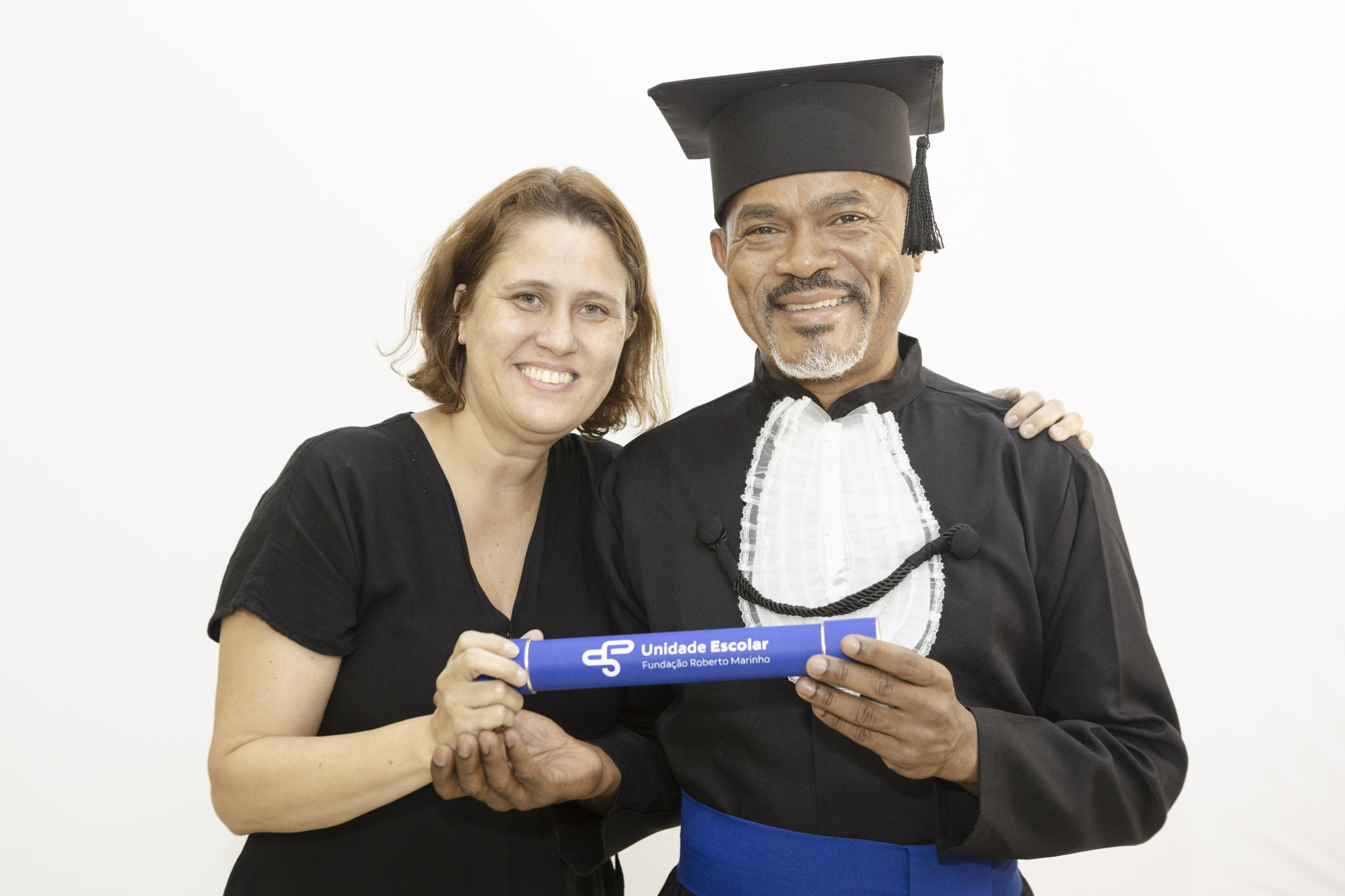 Duas pessoas posam juntas diante de um fundo branco. À direita, um homem sorri e veste beca preta de formatura, com faixa azul na cintura, jabô branco no peito e capelo na cabeça. Ele segura um canudo azul com a inscrição “Unidade Escolar – Fundação Roberto Marinho”. À esquerda, uma mulher também sorri, veste uma blusa preta e apoia a mão no braço do homem, ajudando a segurar o canudo. A imagem transmite proximidade, orgulho e celebração de uma conquista acadêmica.