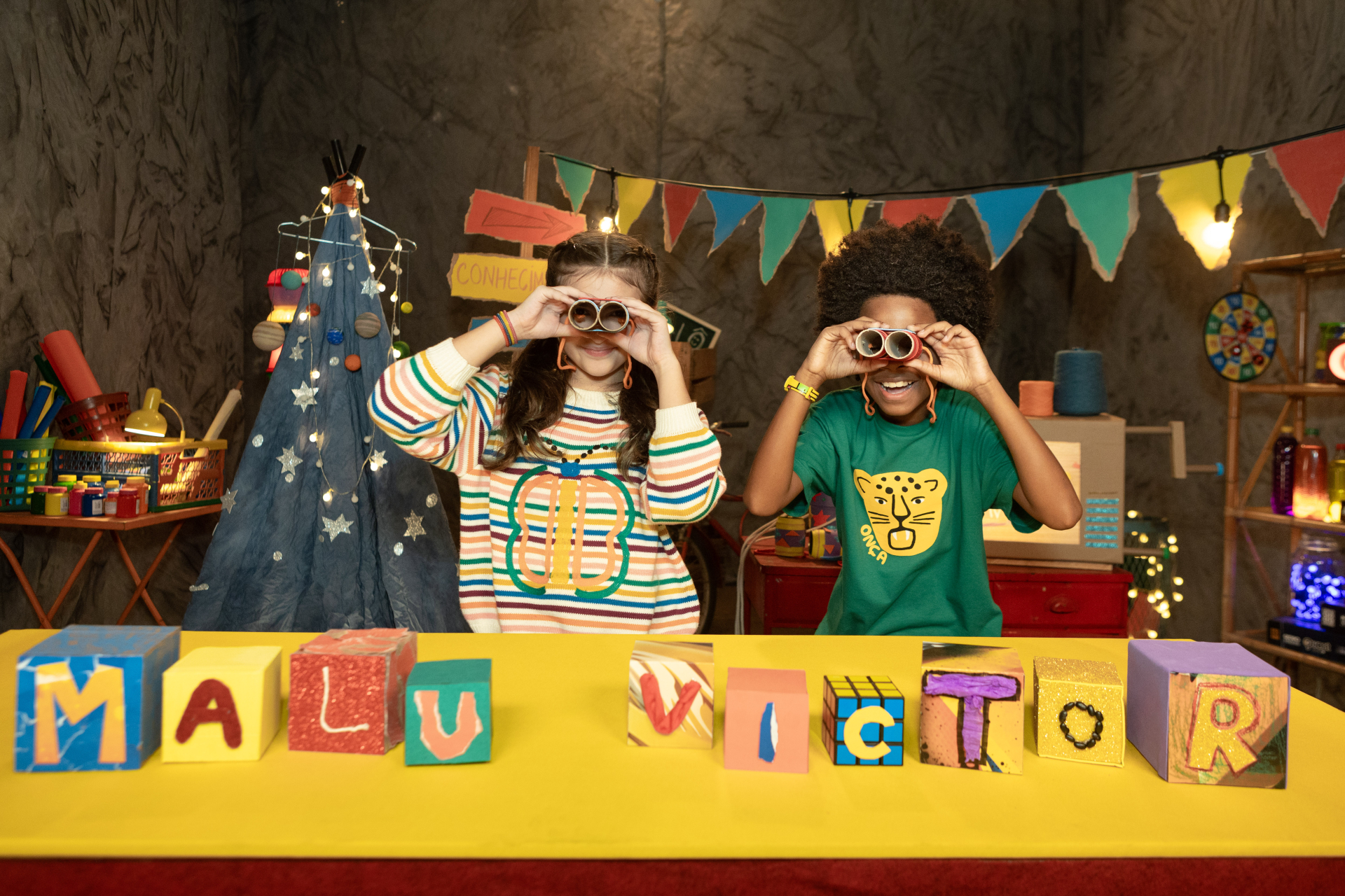 Duas crianças, uma menina branca e um menino negro, estão atrás de uma mesa amarela, sorrindo e olhando pela lente de binóculos feitos à mão. Sobre a mesa, blocos coloridos formam o nome “MALU VICTOR”. O ambiente é lúdico, com bandeirinhas, brinquedos e objetos artesanais ao fundo.