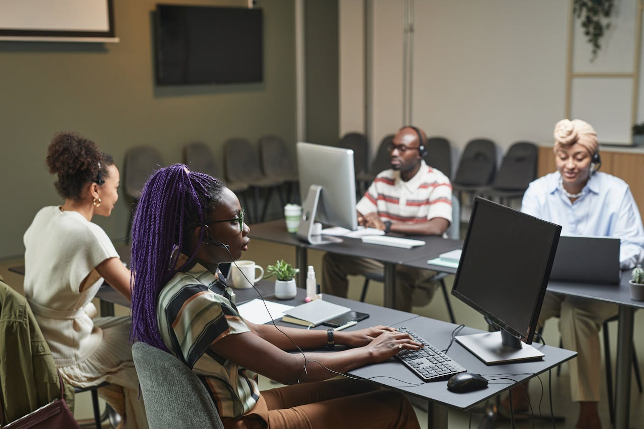 Um grupo de pessoas participa de uma atividade coletiva em um ambiente interno que lembra uma sala de treinamento ou coworking. Sentadas em mesas individuais, elas utilizam computadores e fones de ouvido, concentradas em tarefas digitais ou em uma atividade guiada. A disposição do espaço, os equipamentos e a postura dos participantes sugerem um contexto de capacitação, trabalho colaborativo ou aula prática, marcado por foco, troca e uso de tecnologia.