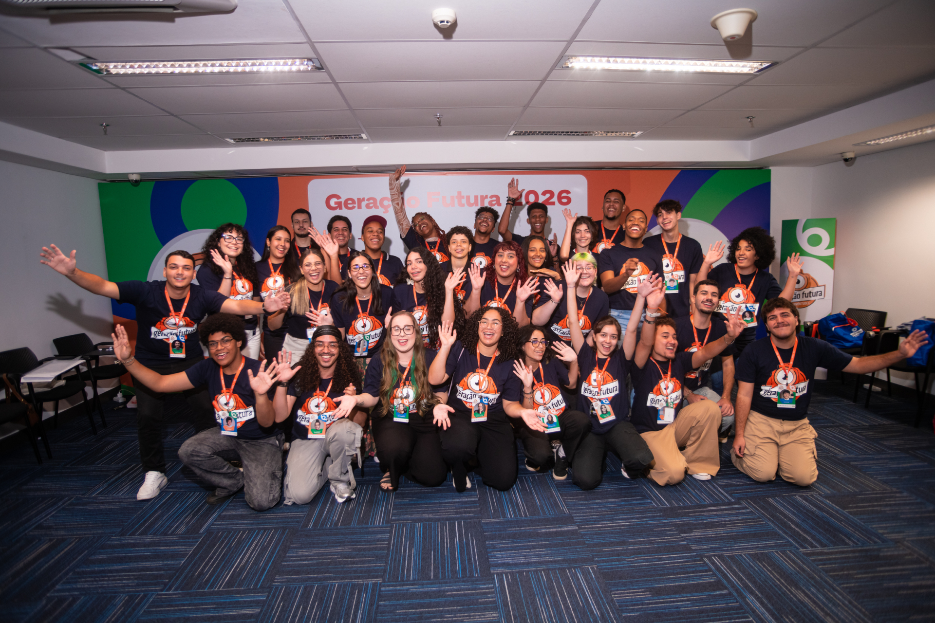 Foto de grupo em ambiente interno, com cerca de 30 jovens reunidos em uma sala ampla, iluminada por luz branca no teto. Todos vestem camisetas azul-marinho com o logotipo laranja do projeto “Geração Futura – Juventudes” e utilizam crachás pendurados em cordões laranja no pescoço.  O grupo está organizado em três fileiras: algumas pessoas sentadas no chão à frente, outras ajoelhadas ou agachadas no centro e o restante em pé atrás.