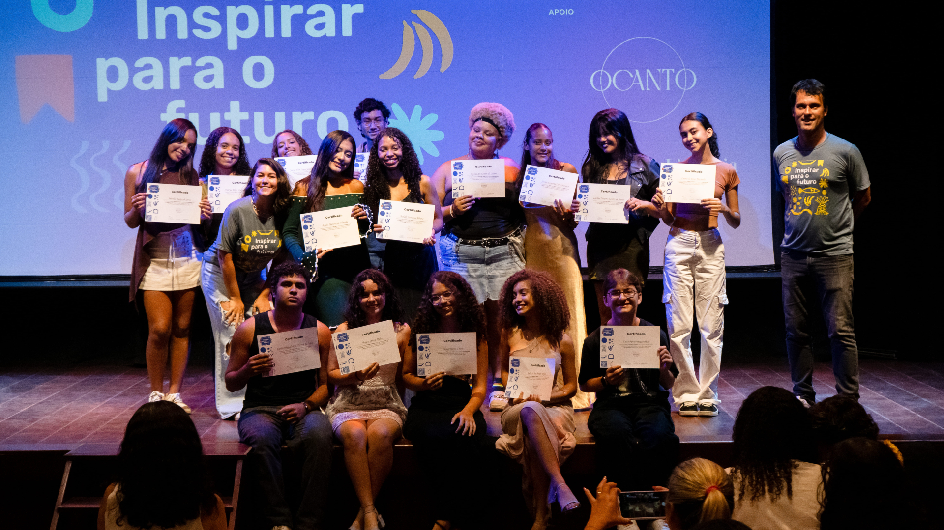 Um grupo de jovens está reunido no palco para uma foto coletiva após o encerramento de uma atividade. Eles seguram certificados e sorriem para a câmera. O grupo está organizado em duas fileiras, com alguns participantes em pé e outros sentados à frente.