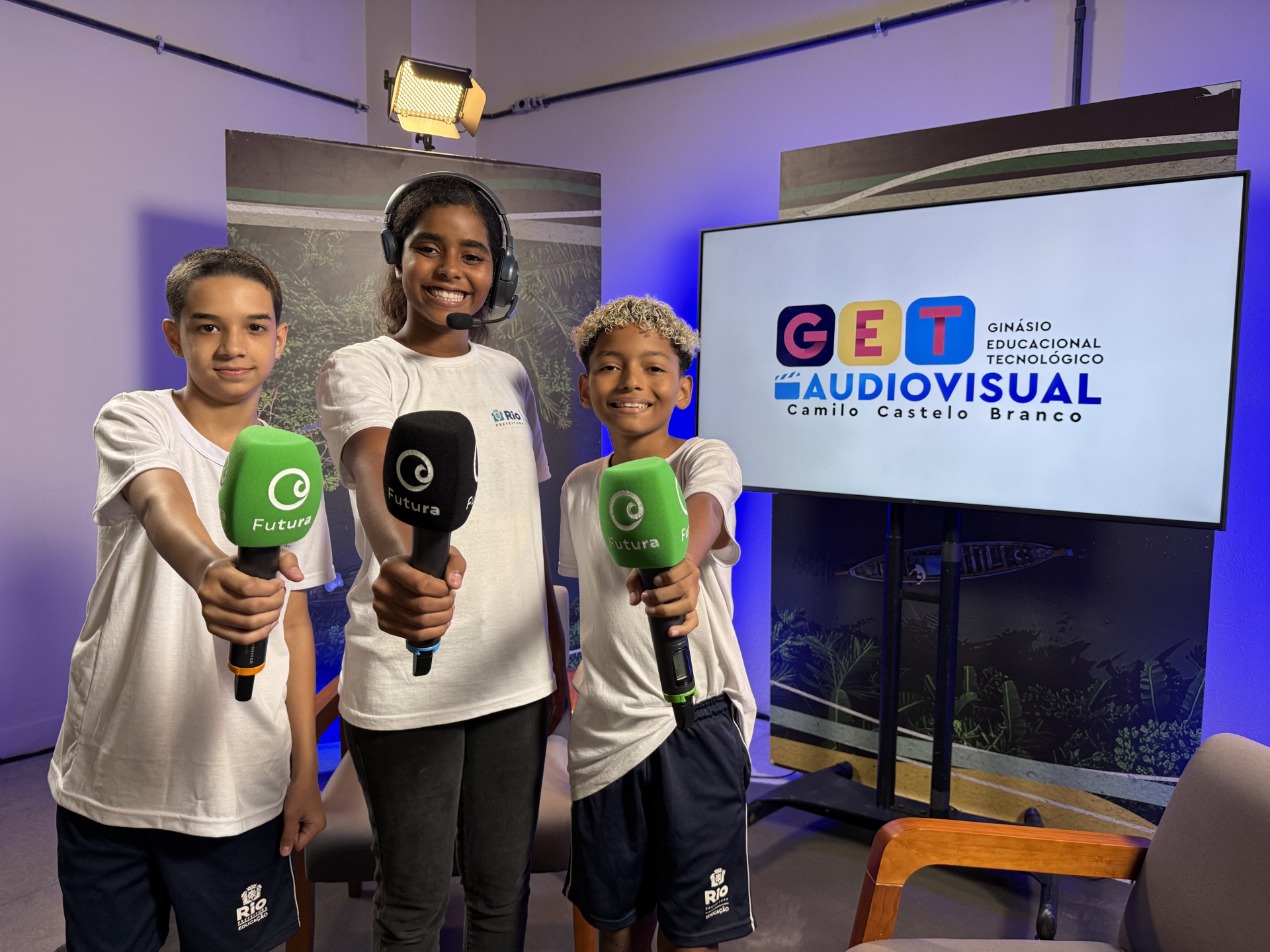 três estudantes aparecem em um estúdio audiovisual segurando microfones com a marca do Canal Futura. Atrás deles, um monitor exibe a identidade do GET Audiovisual – Ginásio Educacional Tecnológico Camilo Castelo Branco, sugerindo um ambiente de aprendizado prático em comunicação e produção de conteúdo.