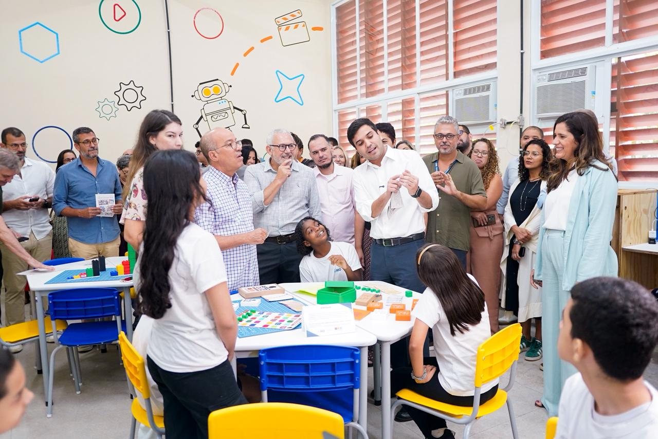 Na última imagem, autoridades e convidados visitam uma sala de aula onde estudantes apresentam atividades em mesas com jogos e materiais pedagógicos. Adultos observam atentamente a demonstração dos alunos, em um ambiente colorido e colaborativo que evidencia práticas de aprendizagem ativa.