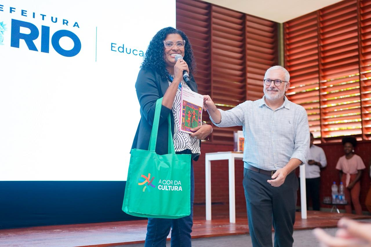uma mulher segura um microfone e apresenta materiais do programa A Cor da Cultura, enquanto recebe um kit educativo das mãos de um homem. Ao fundo, aparece a identidade visual da Prefeitura do Rio, indicando o caráter institucional da atividade.