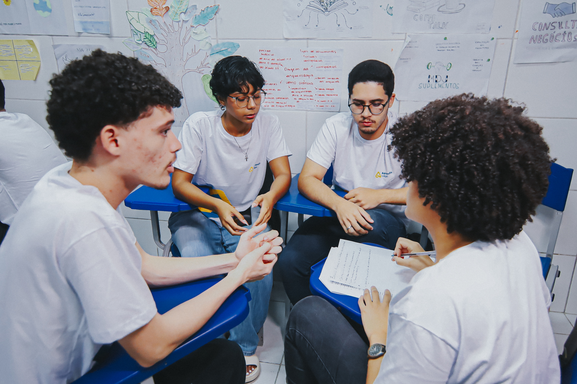 Quatro jovens estão sentados em círculo em uma sala de aula, usando camisetas brancas com um pequeno logotipo. Eles conversam de forma concentrada, em um momento de discussão em grupo. Um deles gesticula com as mãos enquanto fala, outro observa atentamente, e uma jovem segura um caderno, anotando pontos da conversa.
