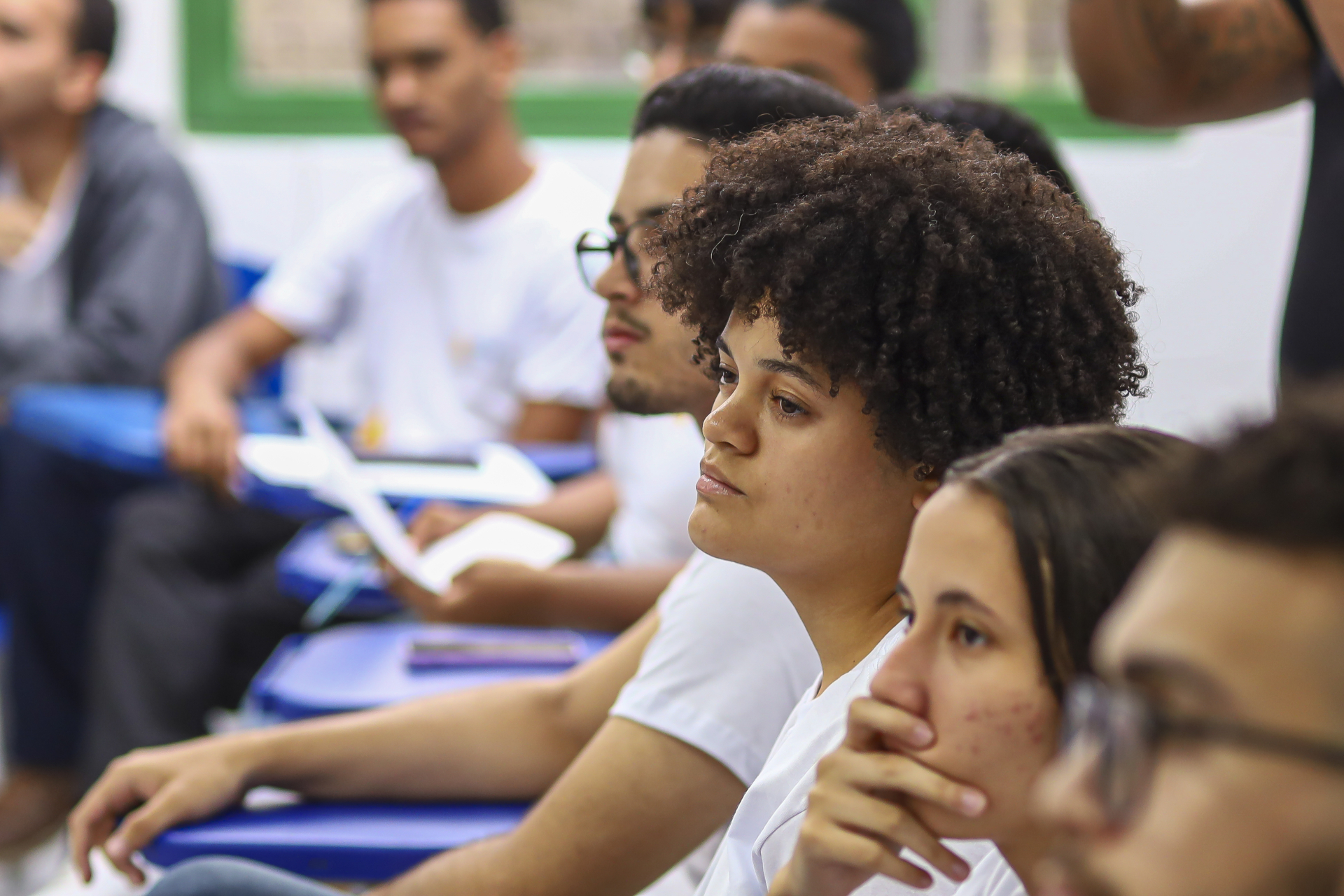 Close em estudantes sentados em sala de aula, olhando à frente com atenção. Em primeiro plano, uma jovem com cabelo cacheado observa concentrada, enquanto outros colegas aparecem ao fundo, também atentos. O ambiente sugere um momento de escuta, como uma explicação ou apresentação.