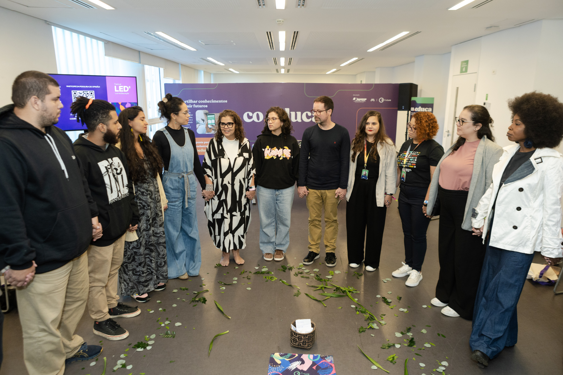Grupo de cerca de dez pessoas forma um círculo de mãos dadas em uma sala de evento, com folhas espalhadas no chão e um pequeno cesto ao centro. Ao fundo, um painel roxo do Festival LED reforça o clima de acolhimento e troca coletiva.