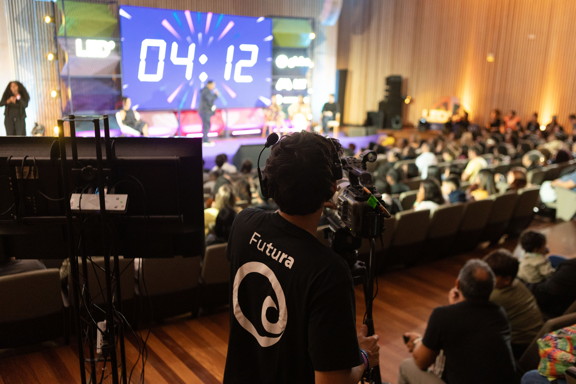 Auditório cheio durante uma programação do Festival LED, visto do fundo da sala. Em primeiro plano, um cinegrafista da equipe do Canal Futura grava o palco iluminado, onde um grande cronômetro digital marca 4 minutos e 12 segundos.