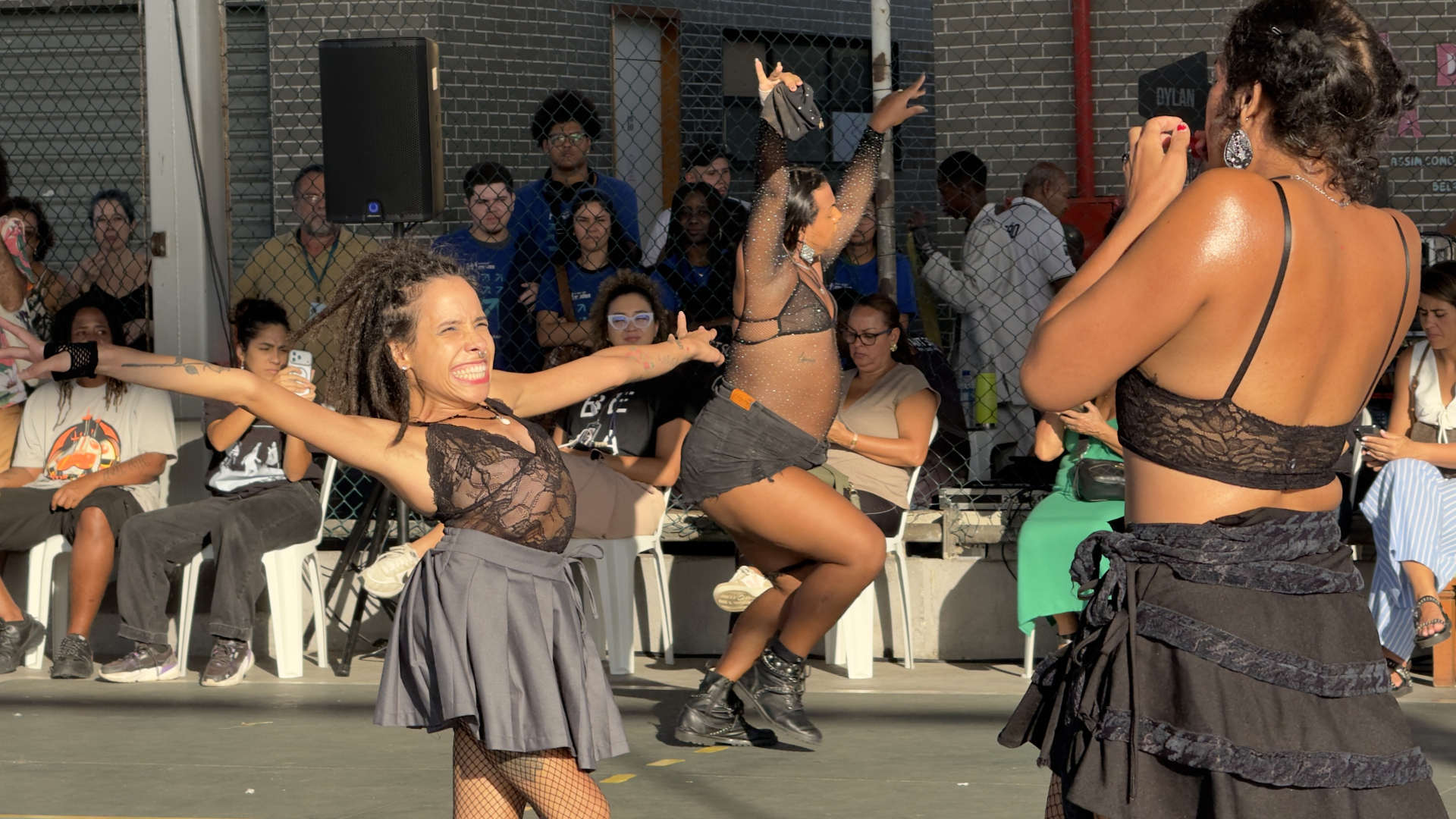 A imagem mostra uma apresentação de dança ao ar livre, com forte energia e expressividade.  No primeiro plano, uma das performers abre os braços e sorri intensamente, transmitindo alegria e entrega ao movimento. Ao redor, outras artistas dançam com gestos amplos e coreografias marcantes, vestindo figurinos predominantemente pretos que destacam o corpo e a dinâmica da cena.