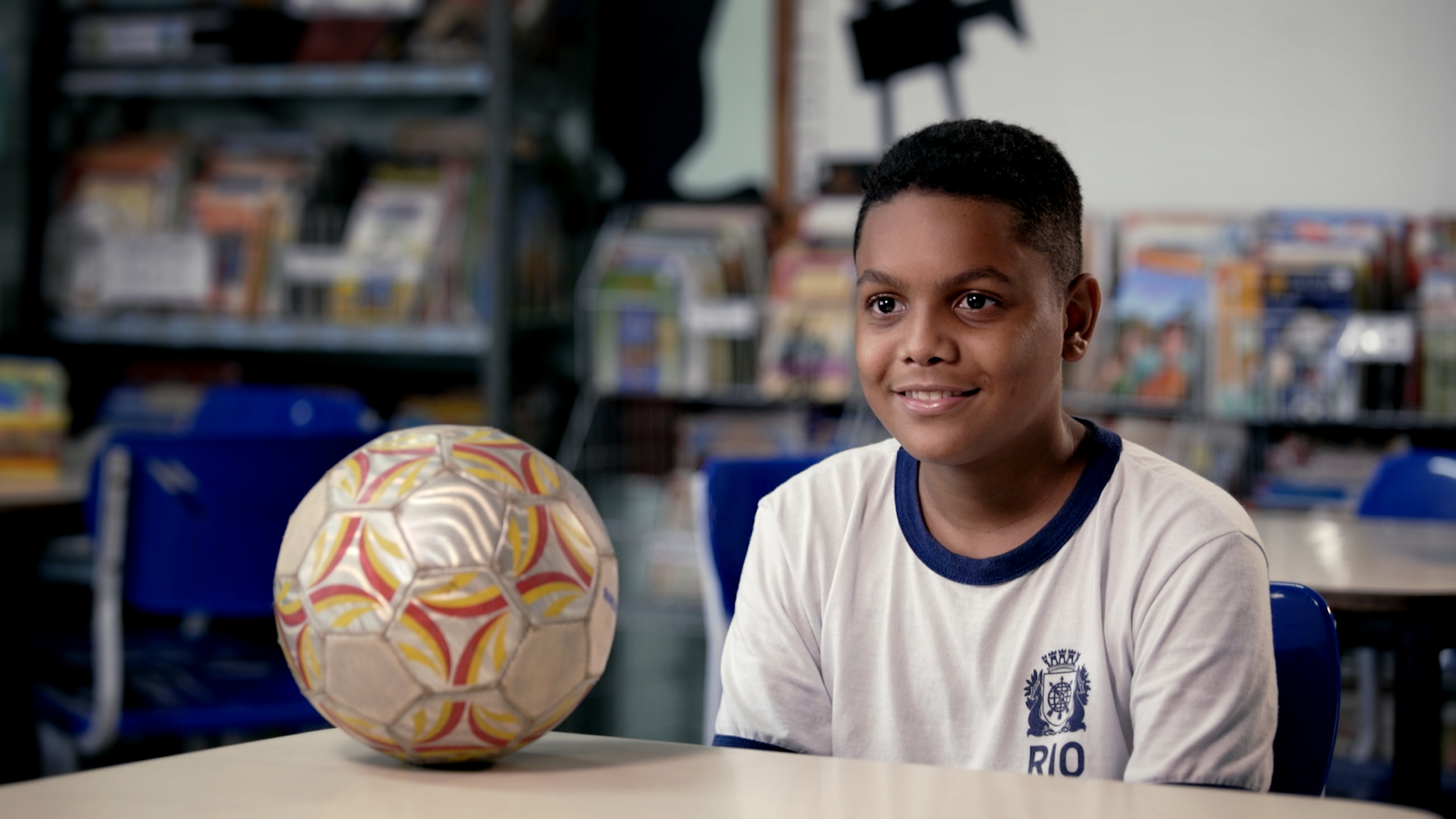 um menino de uniforme escolar branco está sentado em uma biblioteca ou sala de leitura, sorrindo diante de uma bola de futebol apoiada sobre a mesa. Ao fundo, aparecem estantes com livros e cadeiras azuis, reforçando o contexto escolar e educativo.