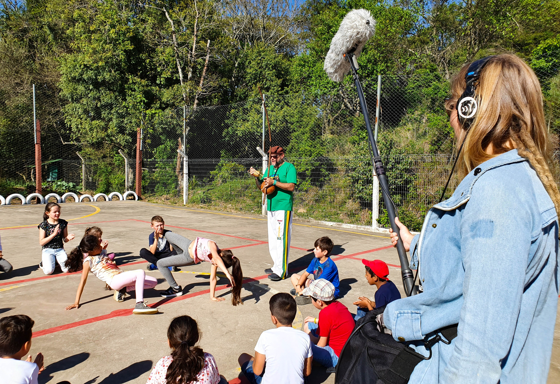 Na primeira imagem, em uma quadra ao ar livre cercada por árvores, crianças participam de uma atividade cultural em roda. No centro, um educador conduz a dinâmica enquanto duas meninas fazem um movimento corporal no chão. À direita, uma profissional com fones de ouvido segura um microfone boom, registrando o som da cena.