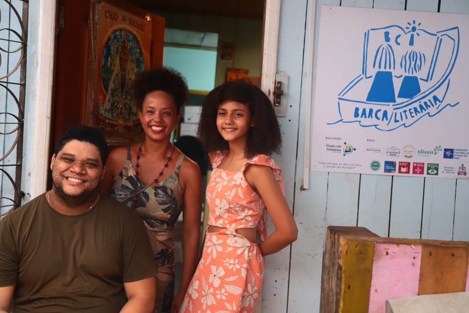 Três pessoas sorriem em frente à entrada de um espaço comunitário. À esquerda, um homem está sentado; ao centro, uma mulher em pé sorri; à direita, uma menina também em pé posa para a foto. Ao lado delas, há um cartaz com a ilustração de um livro aberto sobre um barco e o nome “Barca Literária”, indicando um projeto ligado à leitura e à literatura.