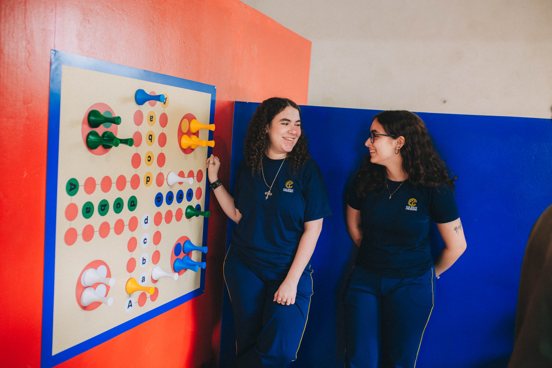 Na primeira imagem, duas estudantes de uniforme azul estão em um ambiente escolar, sorrindo ao lado de um jogo educativo de parede com peças coloridas e letras. O espaço tem paredes em tons de vermelho e azul, criando um ambiente lúdico e interativo de aprendizagem.