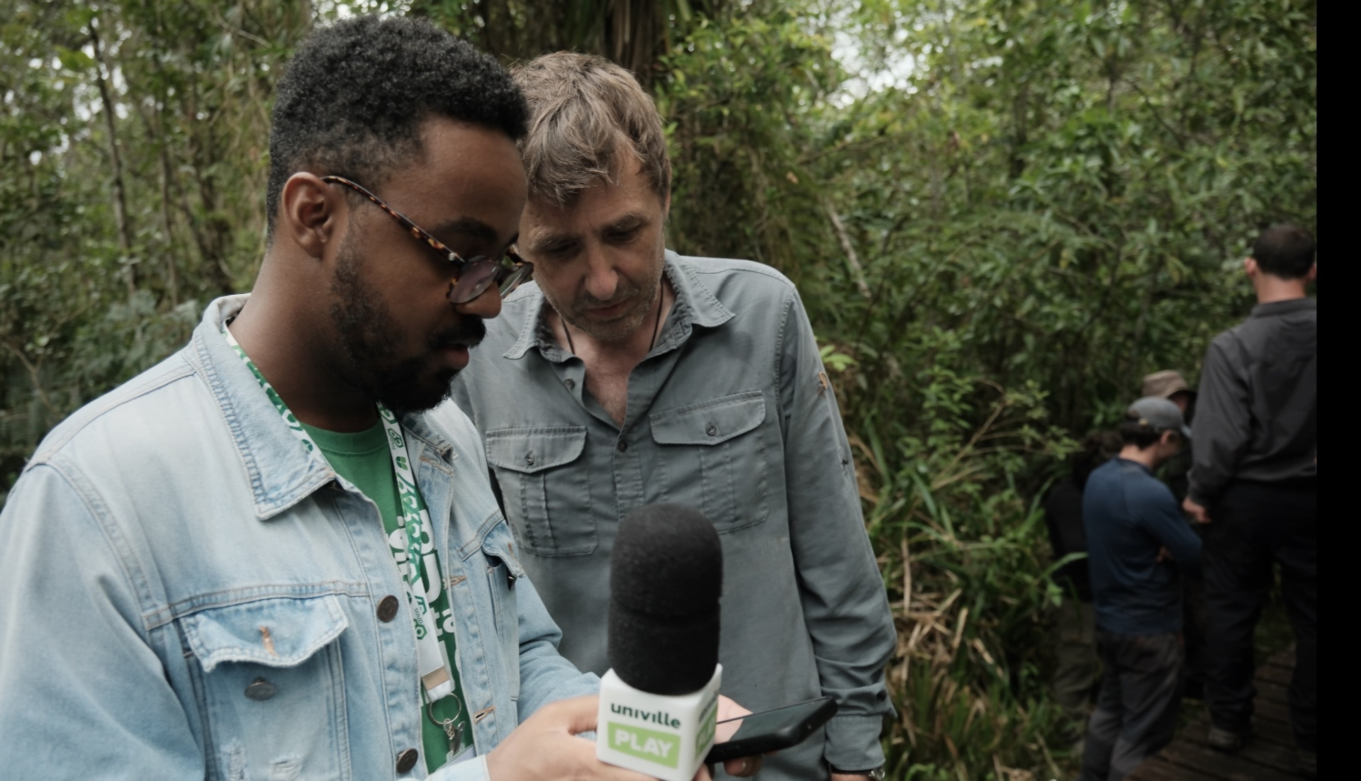 Duas pessoas aparecem no primeiro plano. Um deles segura um microfone, sugerindo uma entrevista. Eles olham algo que o repórter está segurando. 