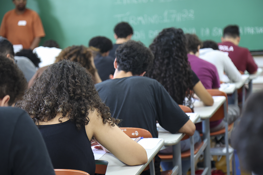 Sala de aula vista por trás, com vários estudantes sentados em fileiras, de cabeça baixa, escrevendo durante uma prova. À frente, um quadro verde e um professor parcialmente visível.