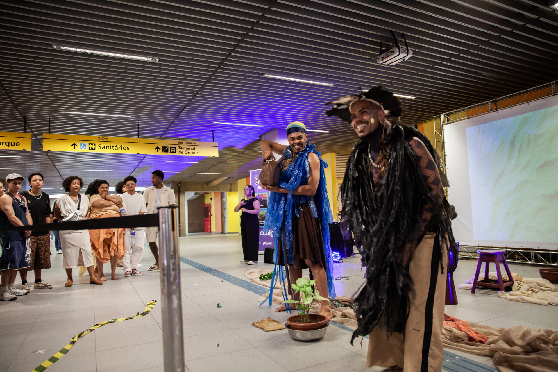 Dois performers estão em destaque: um à frente, sorrindo, com um figurino marcante em tons escuros, com elementos que lembram penas ou fibras, e outro logo atrás, também sorridente, vestindo roupas em azul e marrom e segurando um objeto. Ambos usam microfones de cabeça, sugerindo uma performance que combina fala, música ou teatro.