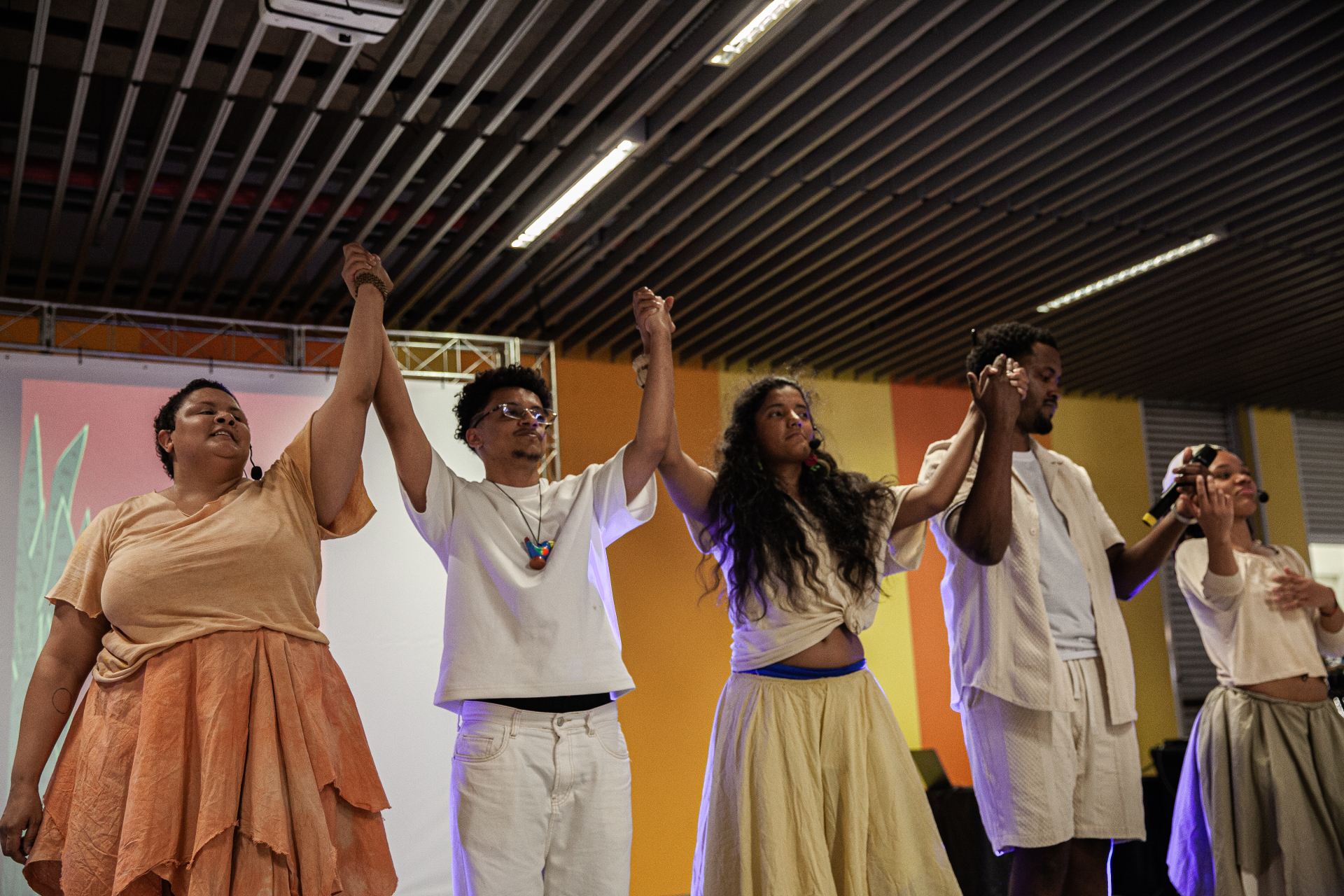 A imagem mostra um grupo de cinco pessoas em um palco, de mãos dadas e com os braços levantados, como se estivessem finalizando uma apresentação e recebendo aplausos.  Elas vestem roupas em tons claros e terrosos, com um estilo leve e fluido, sugerindo uma proposta artística ou cultural. Algumas usam microfones de cabeça, indicando que houve fala ou performance ao vivo. As expressões são sérias ou emocionadas, transmitindo intensidade e conexão com o momento.