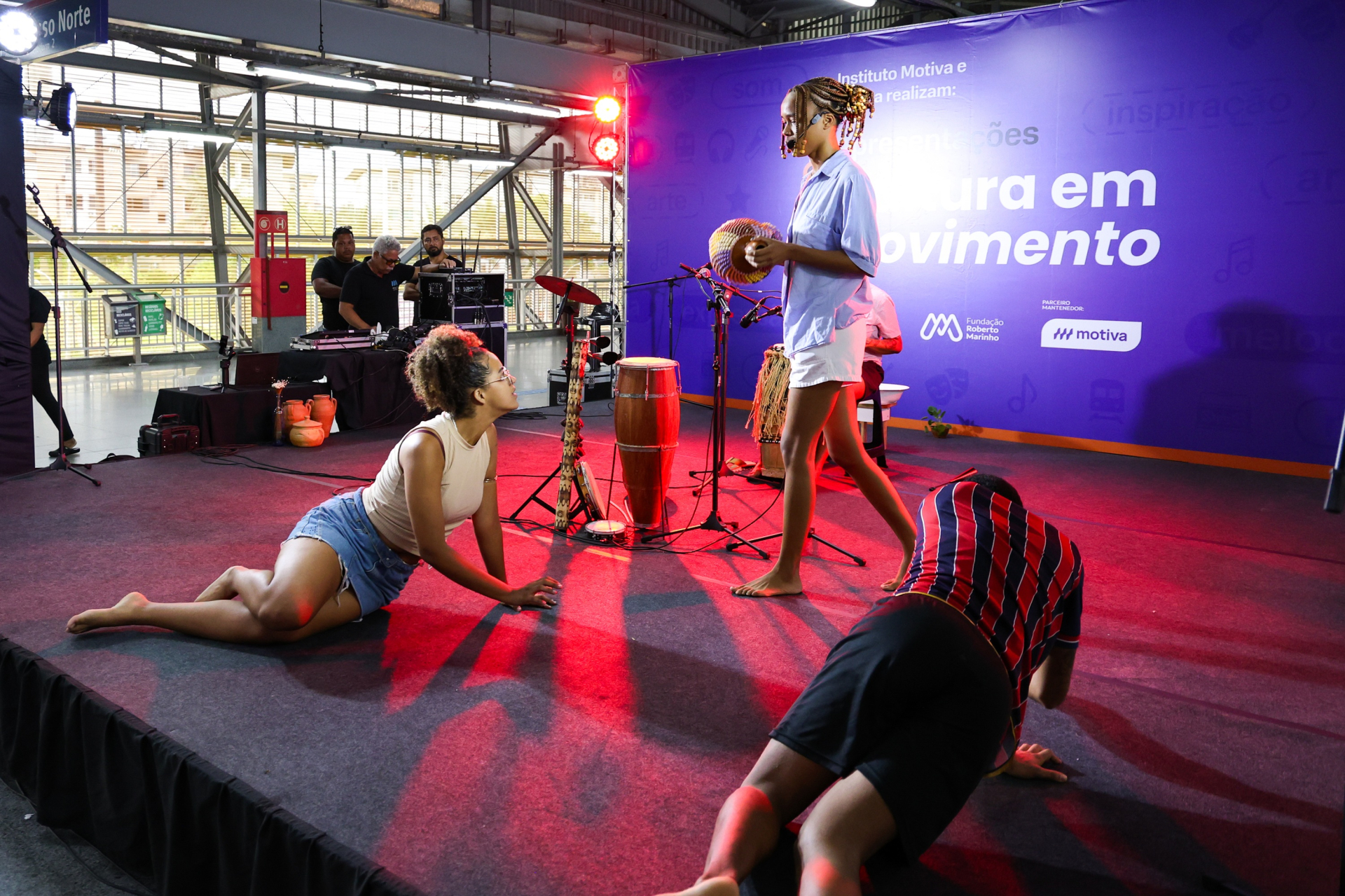 artistas se apresentam no palco com música e expressão corporal. Instrumentos de percussão e iluminação cênica destacam a performance, enquanto os movimentos dos participantes no chão e em pé sugerem uma narrativa artística intensa e sensorial.