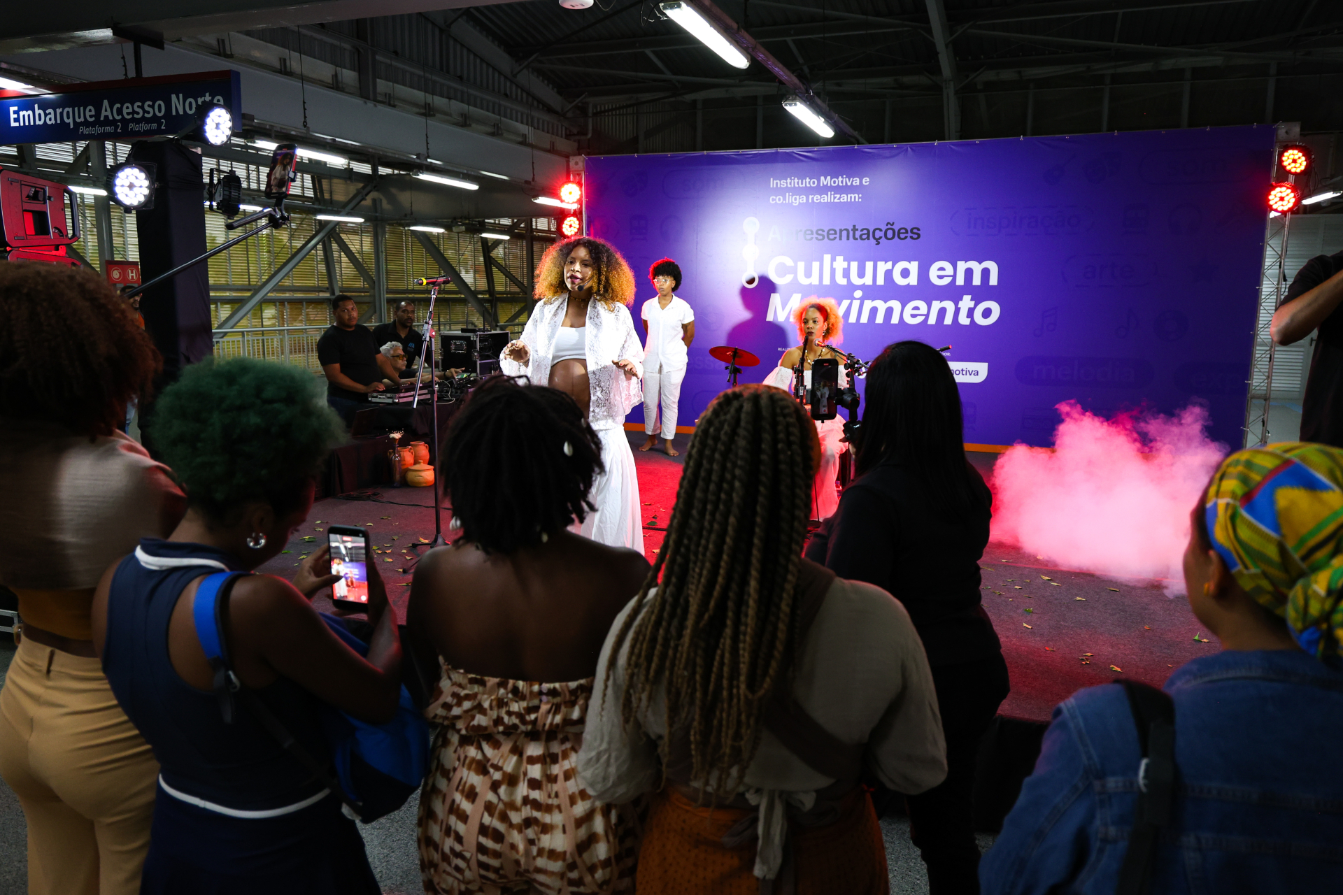 Uma apresentação artística acontece em um espaço que parece ser uma estação ou área coberta de transporte, com estrutura metálica e placas de sinalização ao fundo. No centro, uma mulher se apresenta no palco, usando roupas claras e esvoaçantes, com expressão envolvente e postura de performance. Atrás dela, outros artistas acompanham, incluindo um músico e uma pessoa em pé, compondo a cena.