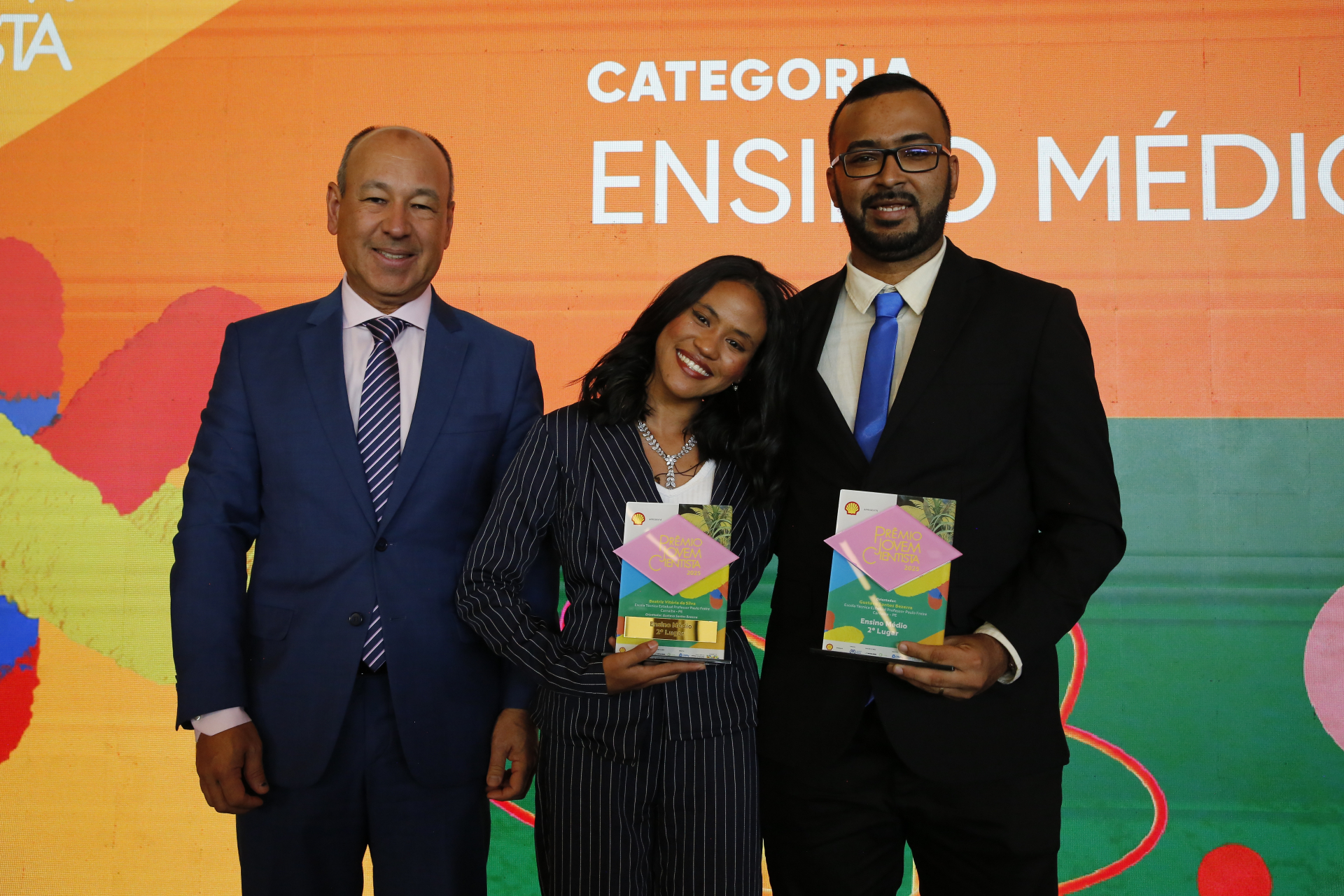 Três pessoas posam lado a lado em um palco, sorrindo para a foto. Ao centro, uma jovem segura um troféu e inclina levemente a cabeça, demonstrando alegria. À direita, um homem também segura um prêmio semelhante. À esquerda, um homem mais velho veste terno e gravata. Ao fundo, há um painel colorido com a inscrição “Categoria Ensino Médio”, indicando uma cerimônia de premiação.