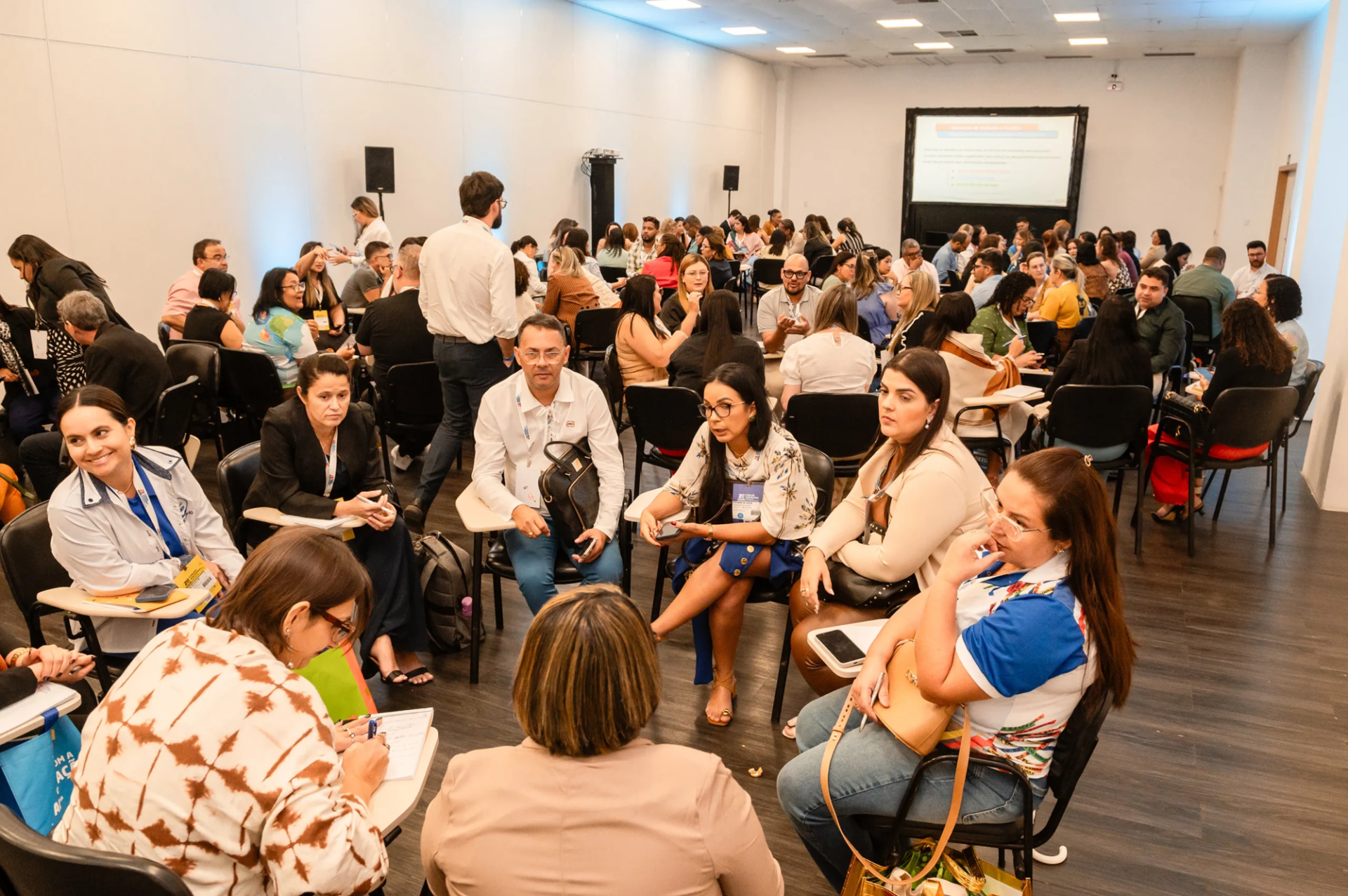 Imagem mostra auditório com cores claras. Há um número grande de pessoas organizadas em alguns círculos. Todos estão sentados e parecem dialogar sobre algum tema. O clima é de integração e aprendizado.