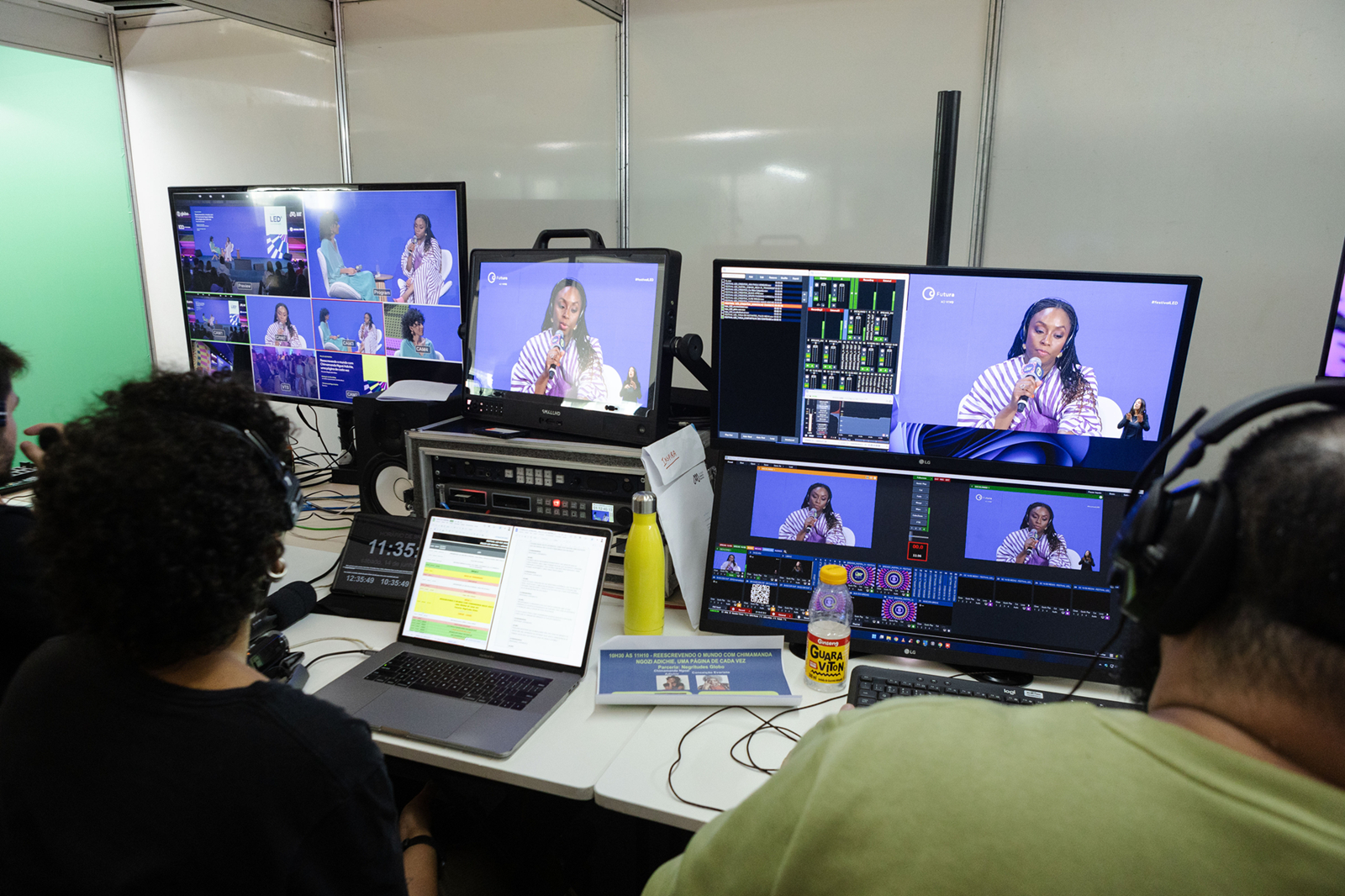 Uma sala de controle de transmissão ao vivo é mostrada cheia de equipamentos eletrônicos e monitores ligados. Duas pessoas aparecem sentadas de costas, usando fones de ouvido e operando computadores e mesas de vídeo. Sobre as mesas há notebooks, teclados, cabos, garrafas e aparelhos de áudio e imagem.  As telas exibem diferentes enquadramentos de uma mulher falando ao microfone em um palco iluminado por luzes azuis e roxas.